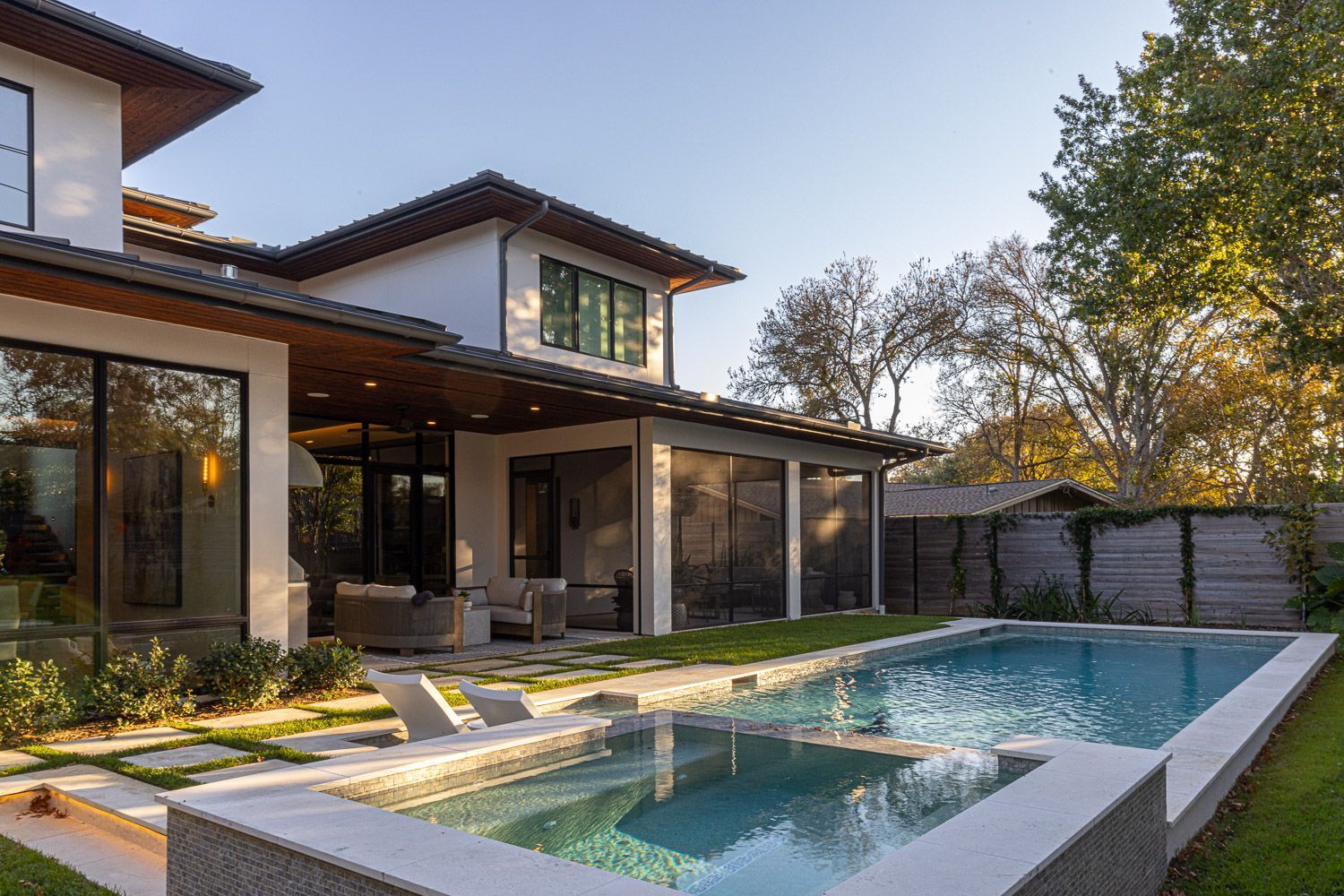 A modern house with a large pool and hot tub in the backyard, set against a background of trees under a clear sky.