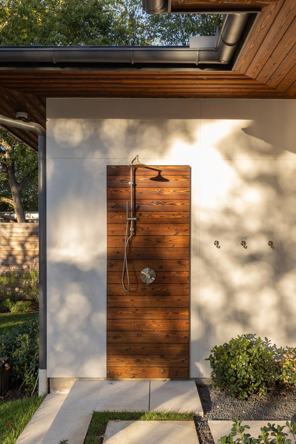 An outdoor shower with a rustic wooden panel mounted on a white wall, featuring a modern metal shower head and fixtures.