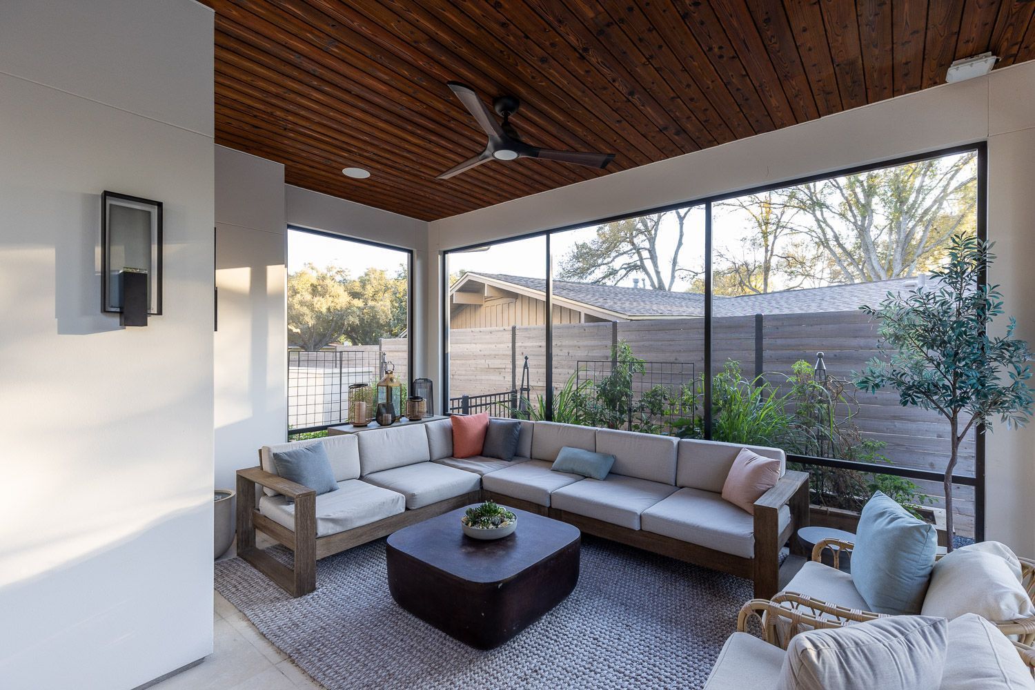 A cozy, modern patio with a wood-slatted ceiling, large floor-to-ceiling windows, and a sectional sofa on a gray rug.