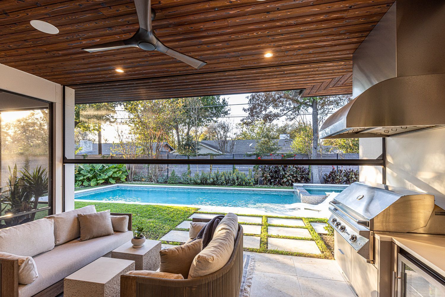 Modern outdoor patio with a swimming pool, lounge seating, and an integrated grill station under a wood-paneled ceiling.