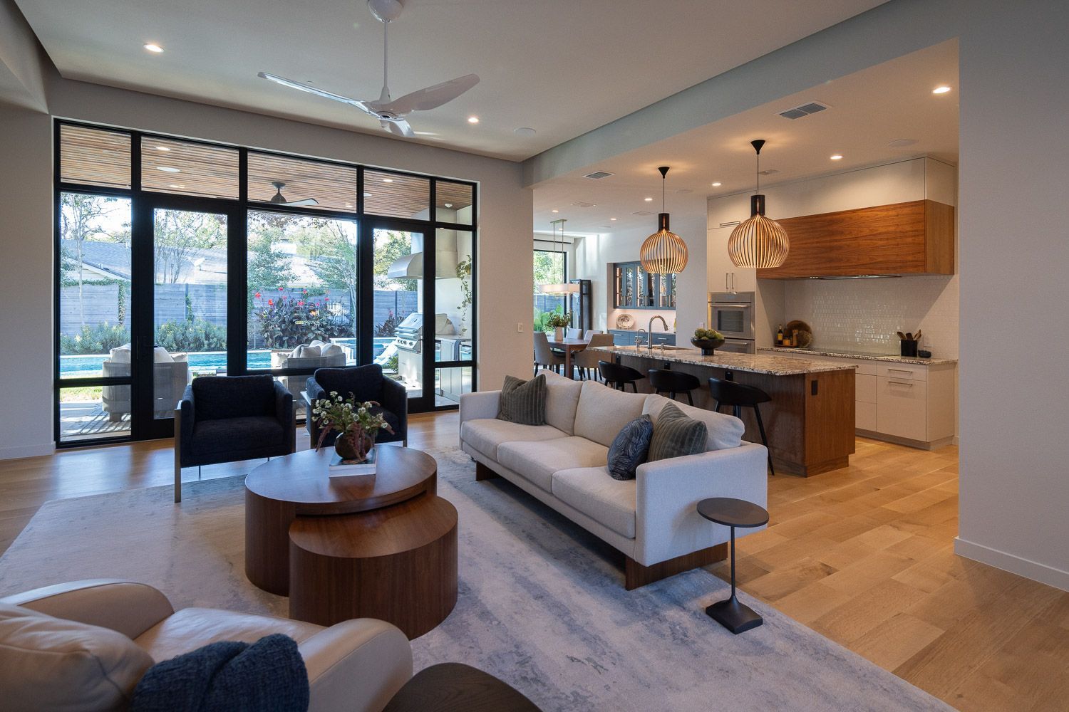 Modern open-concept living area with a white sofa, dark armchairs, wooden coffee tables, and a kitchen with wood cabinets.