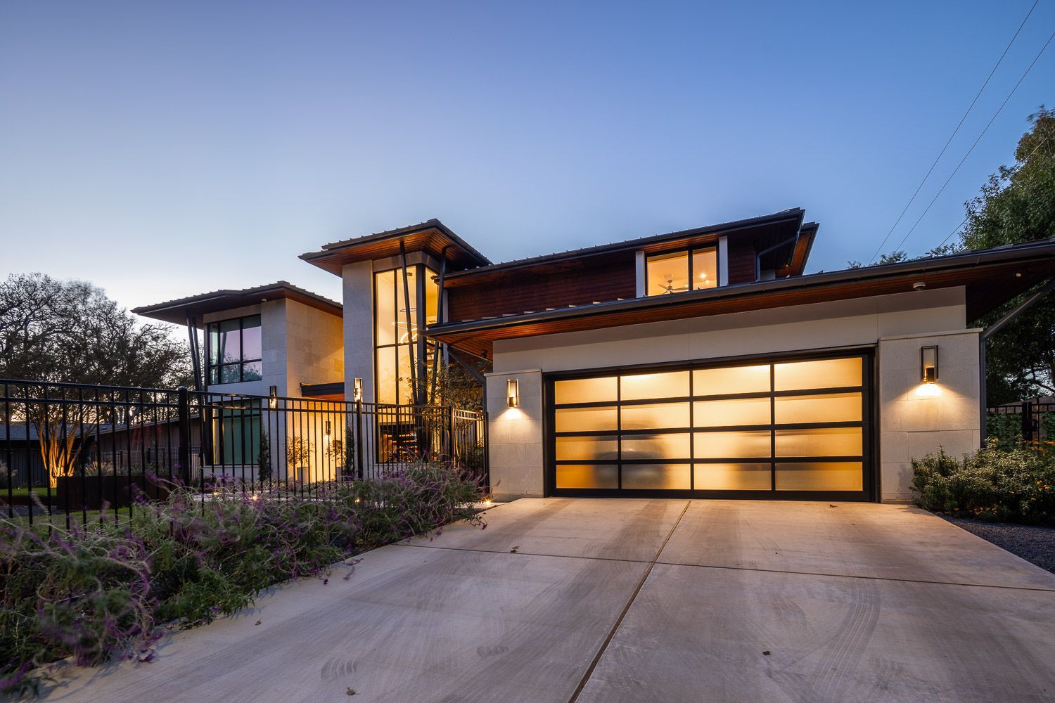A modern, two-story house with a wide, frosted glass garage door, a vertical glass entryway, and warm exterior lighting.
