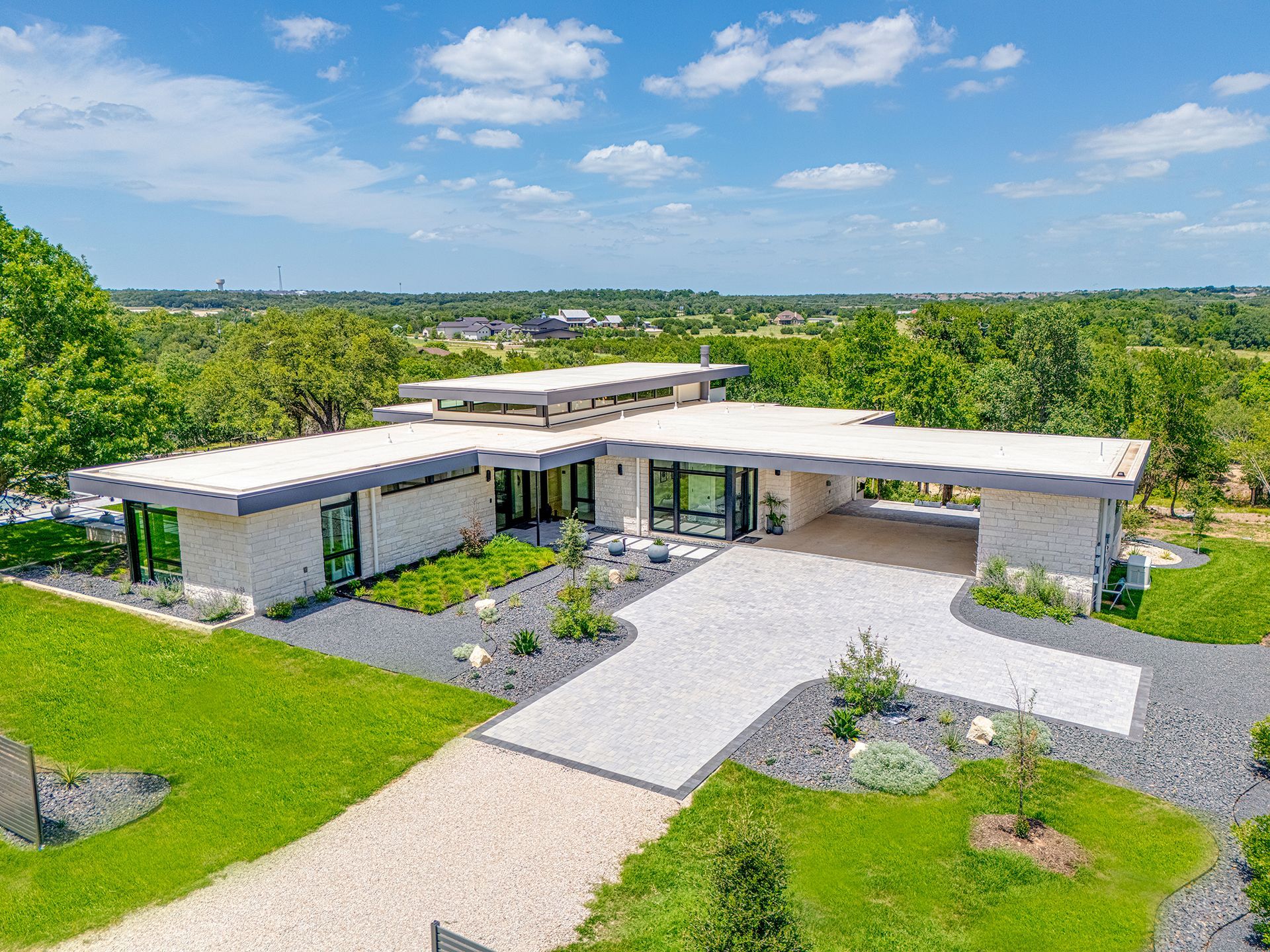 Modern single-story house with flat roofs, large windows, and a gravel driveway amid green landscaping under a blue sky