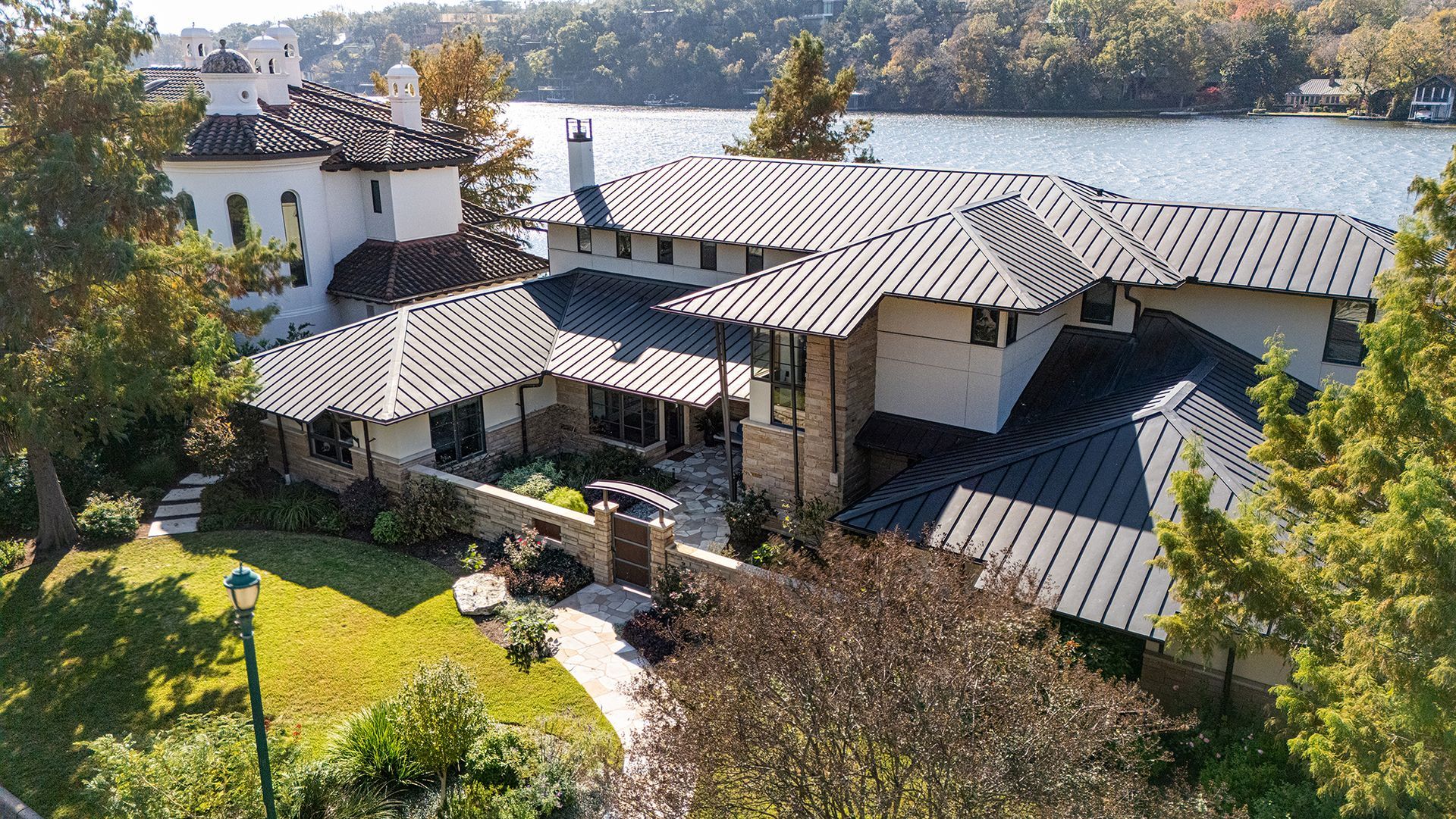 Aerial view of a large white house with a dark metal roof, stone walls, and a lawn, situated next to a body of water.