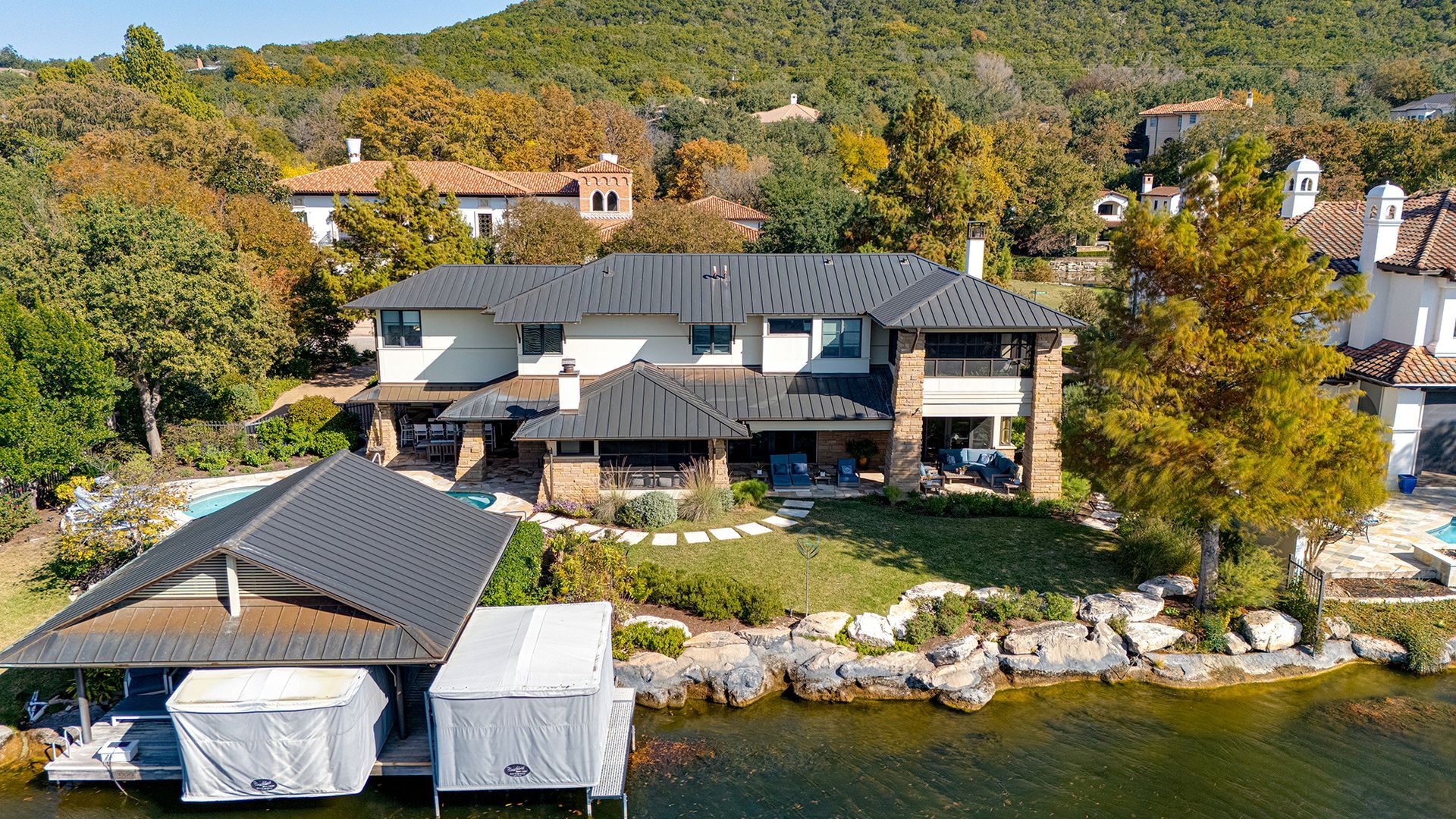 An aerial view of a large white lakeside house with a dark metal roof, stone pillars, and a private covered boat dock.