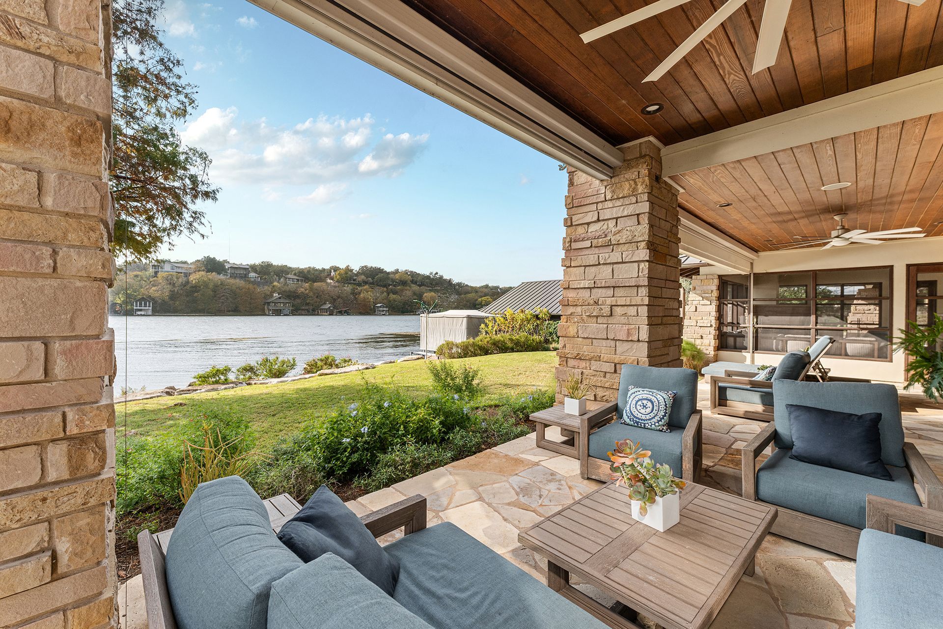 A stone-floored patio with blue outdoor furniture overlooks a grassy lawn and a calm body of water.