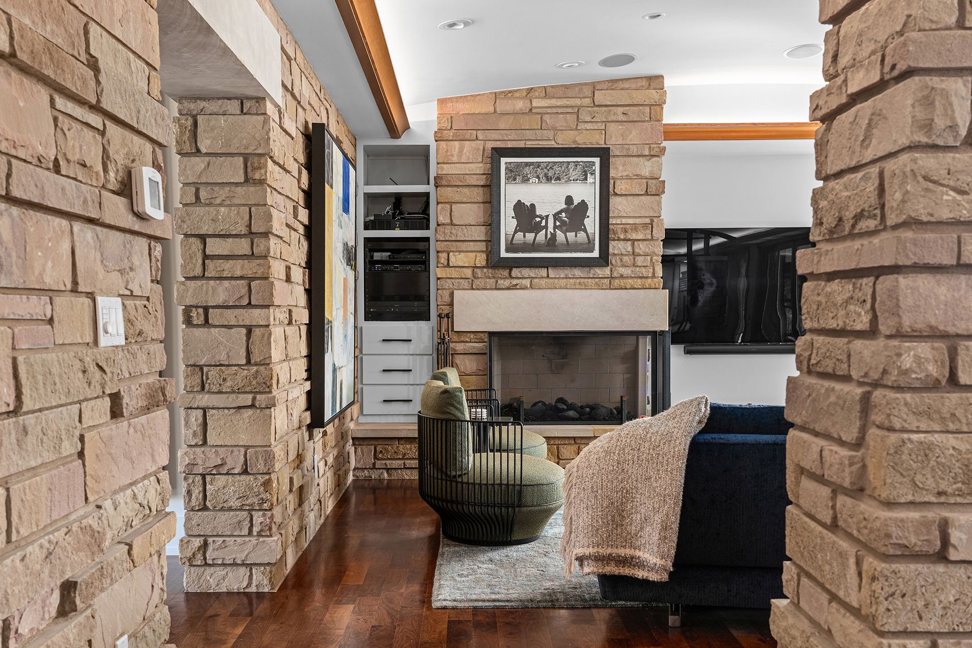 A stone-walled living room with a fireplace, open shelving, a dark swivel chair, and a dark blue sofa with a throw blanket.