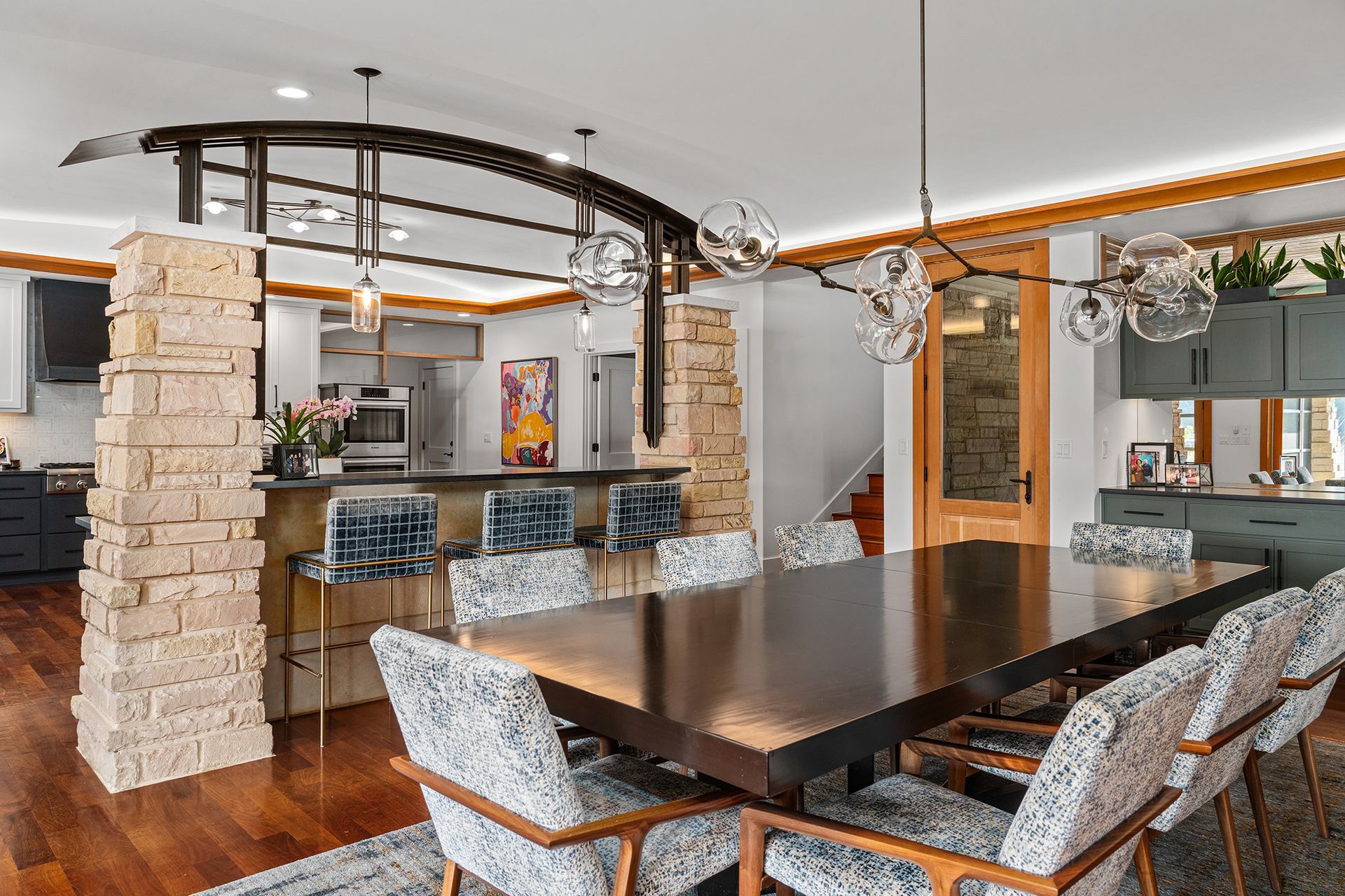 A dining room with a large dark table and patterned chairs, set against a stone pillar and a modern kitchen background.