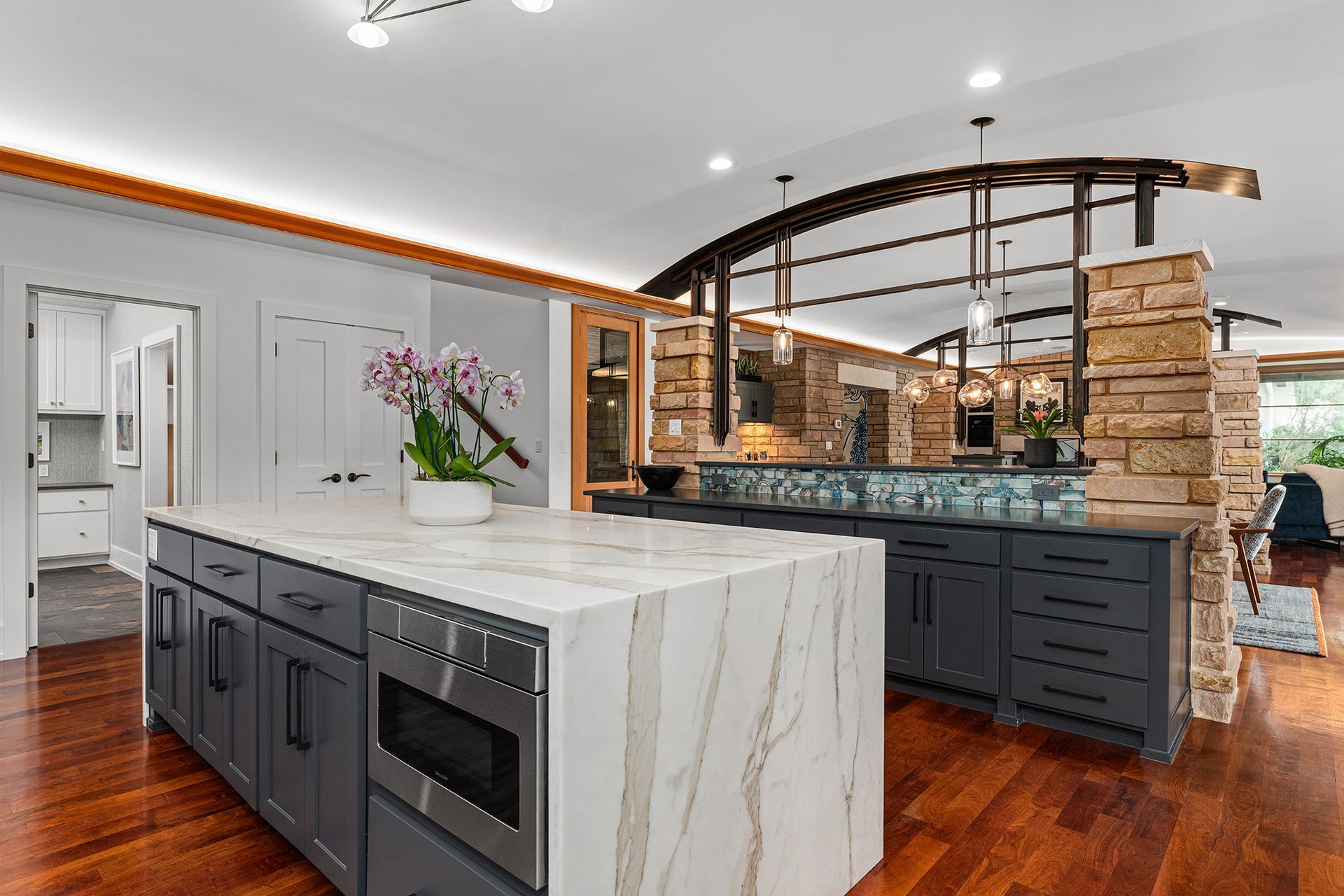 Modern kitchen with a white marble-topped island, dark gray cabinetry, hardwood floors, and a stone-columned breakfast bar.