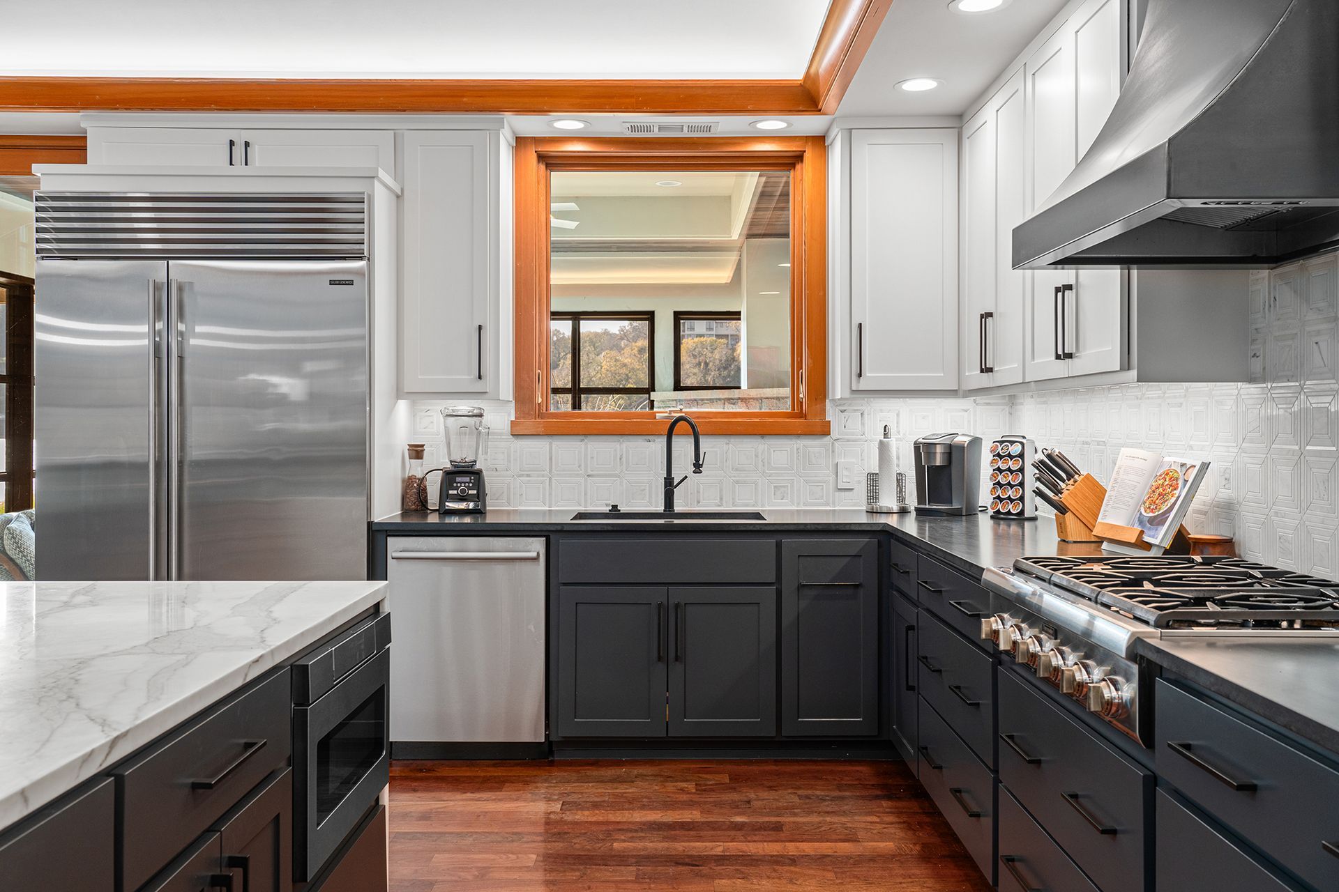 Modern kitchen featuring dark grey cabinets, stainless steel appliances, a white marble island, and a wooden window frame.