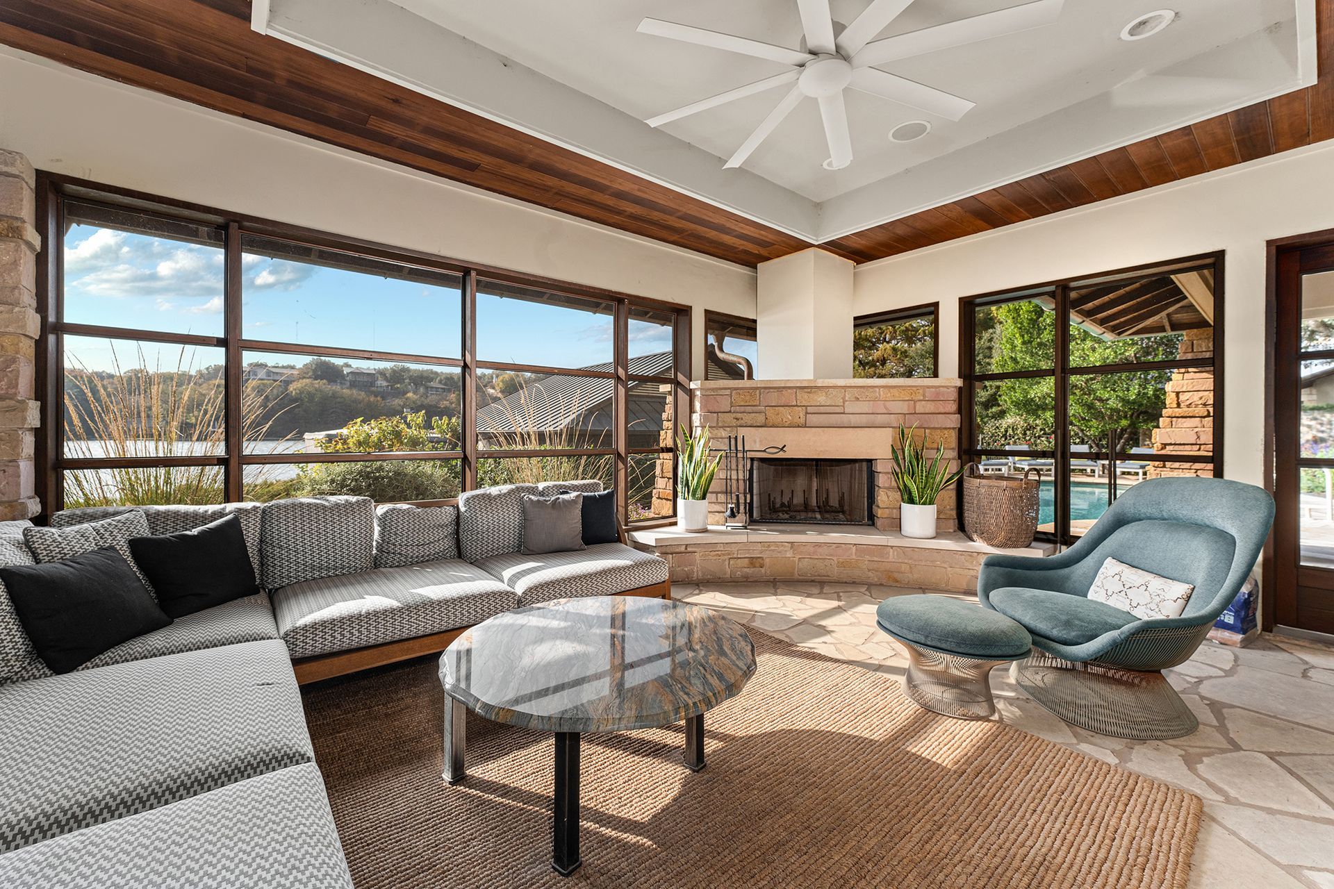 A bright sunroom with a grey sectional, a blue armchair, a round glass coffee table, and a stone fireplace.