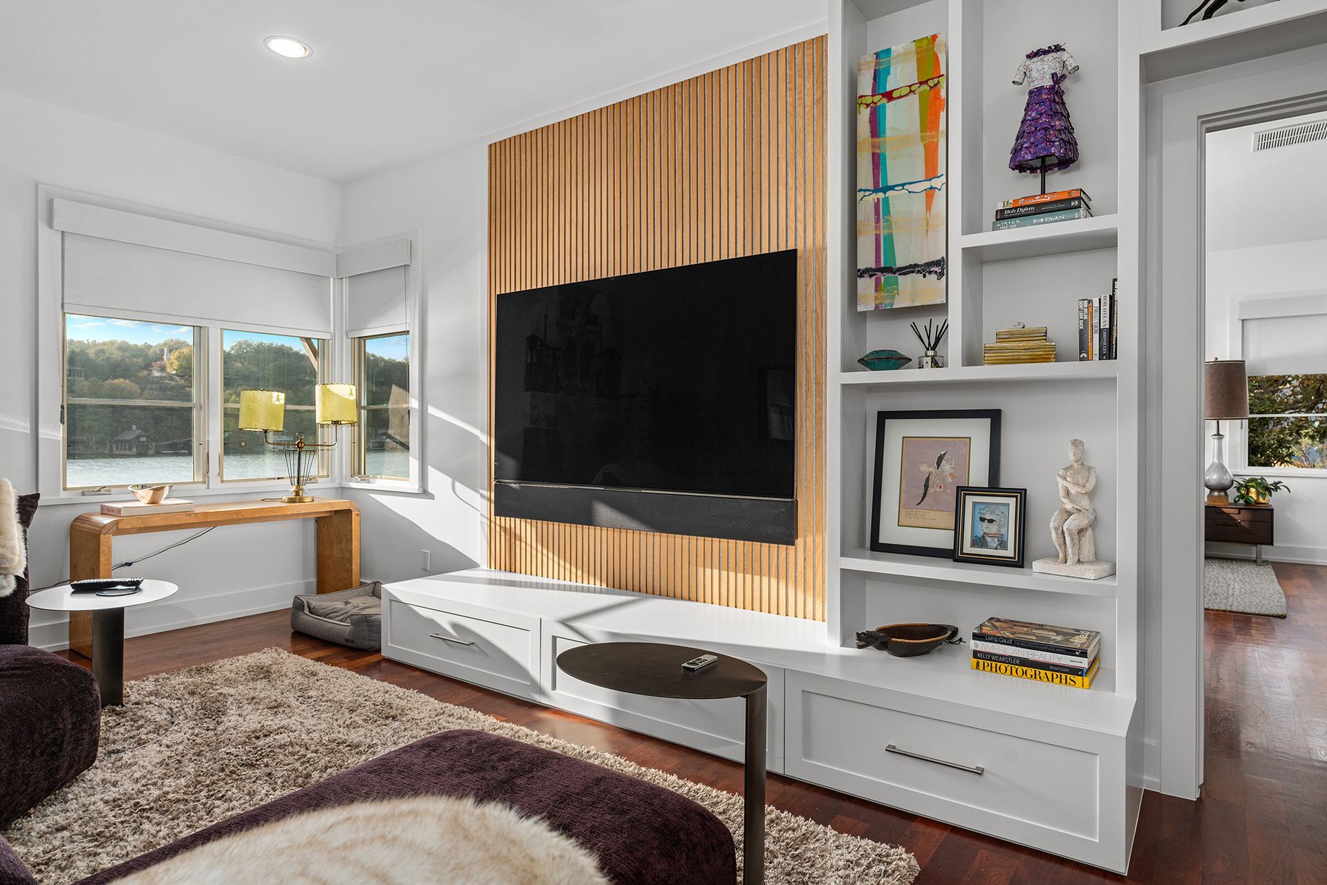 A living room featuring a mounted television on a wood-slat wall, flanked by white built-in shelves and a windowed nook.