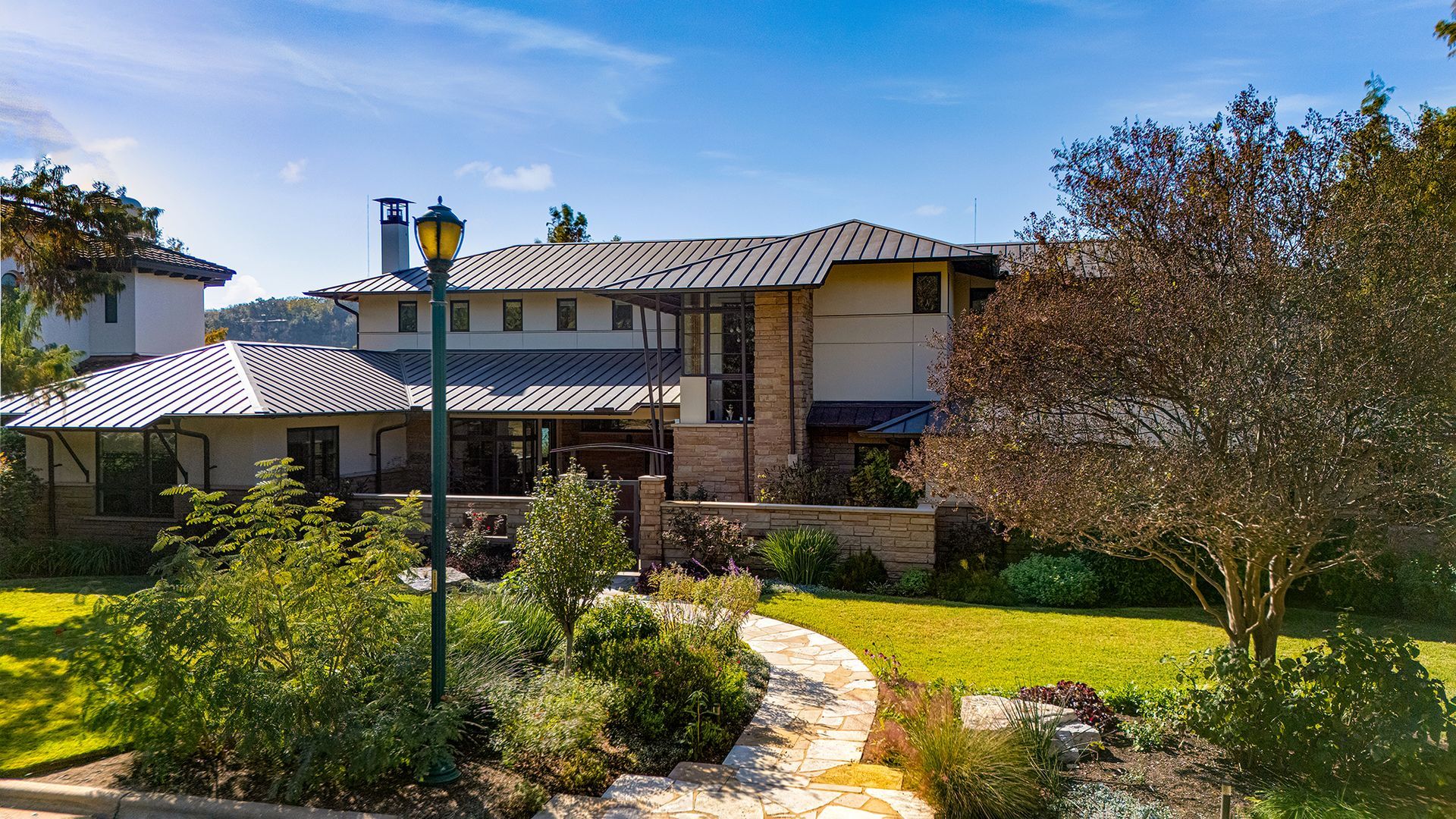 A sprawling two-story modern house with a stone facade, gray roof, and a lush, landscaped garden with a stone path.