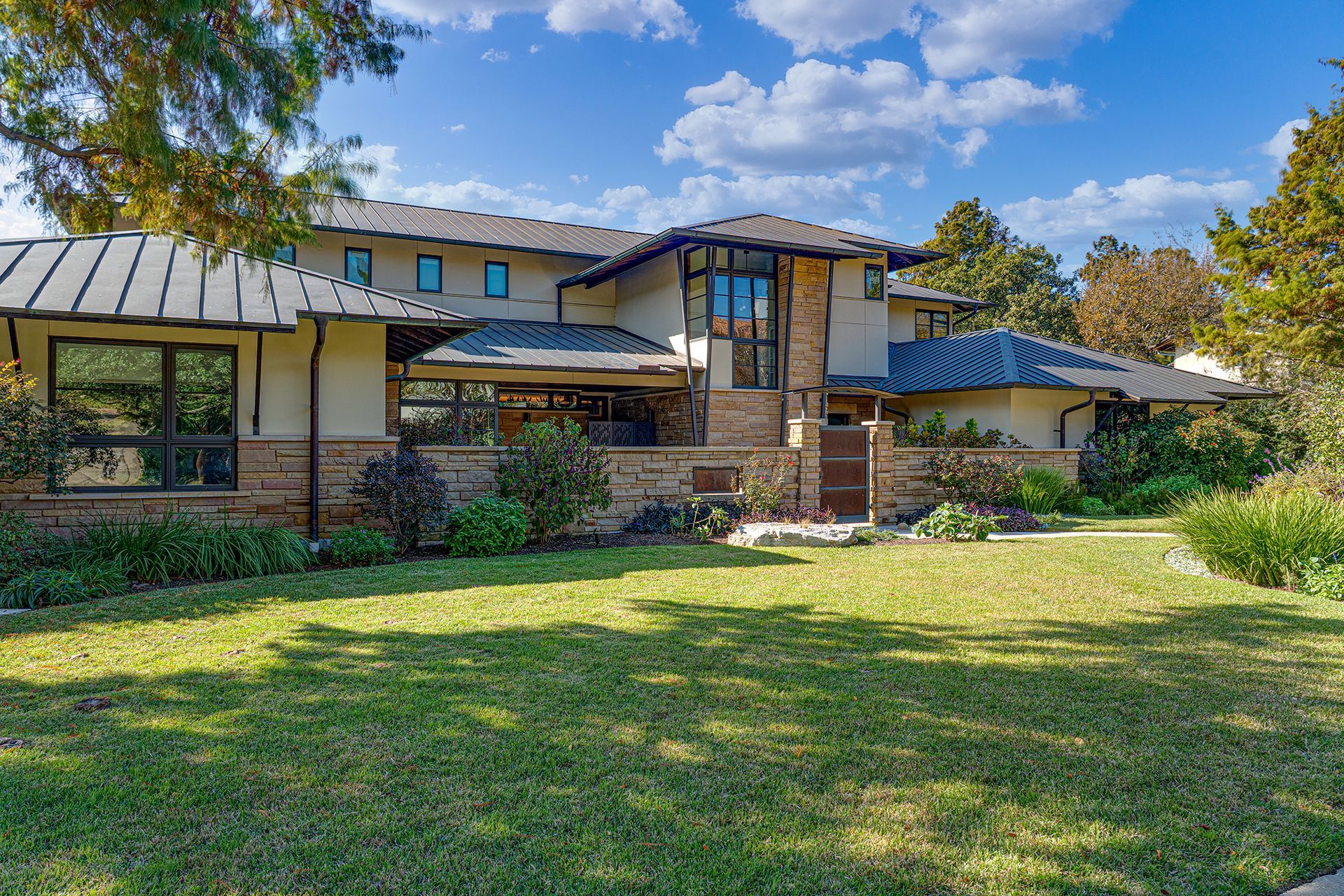 A large modern house with stone accents, multiple roof levels, and a expansive green lawn under a partly cloudy sky.