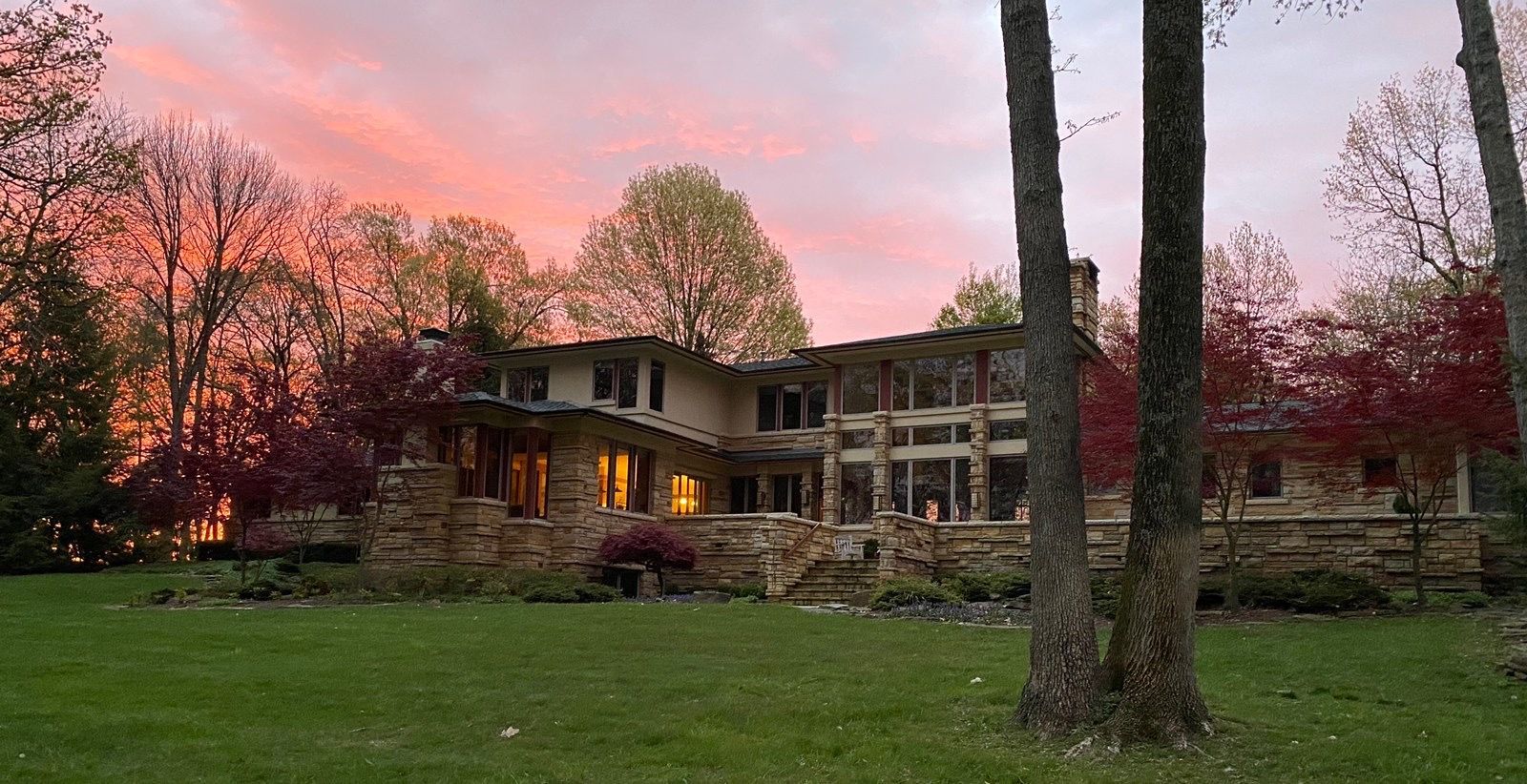 A large house is surrounded by trees and a sunset in the background.