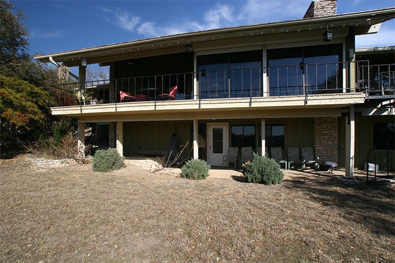 A two-story tan house with a large upper deck, glass doors, and a shaded lower patio area on a dry, grassy lawn.