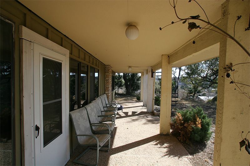 A covered outdoor patio featuring a row of chairs along a white door, with tan walls and concrete flooring.
