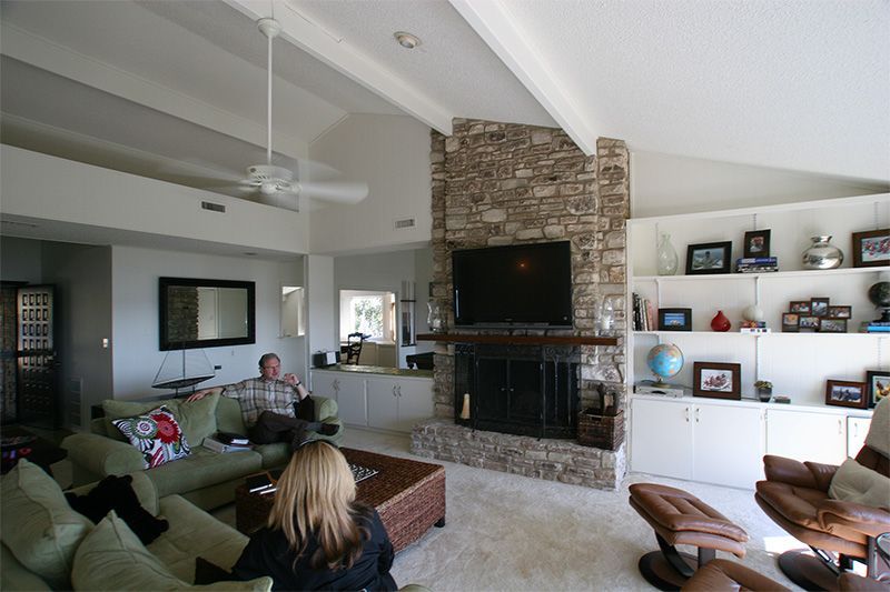 A living room with a stone fireplace, television, built-in shelving, green sofas, and two people sitting inside.