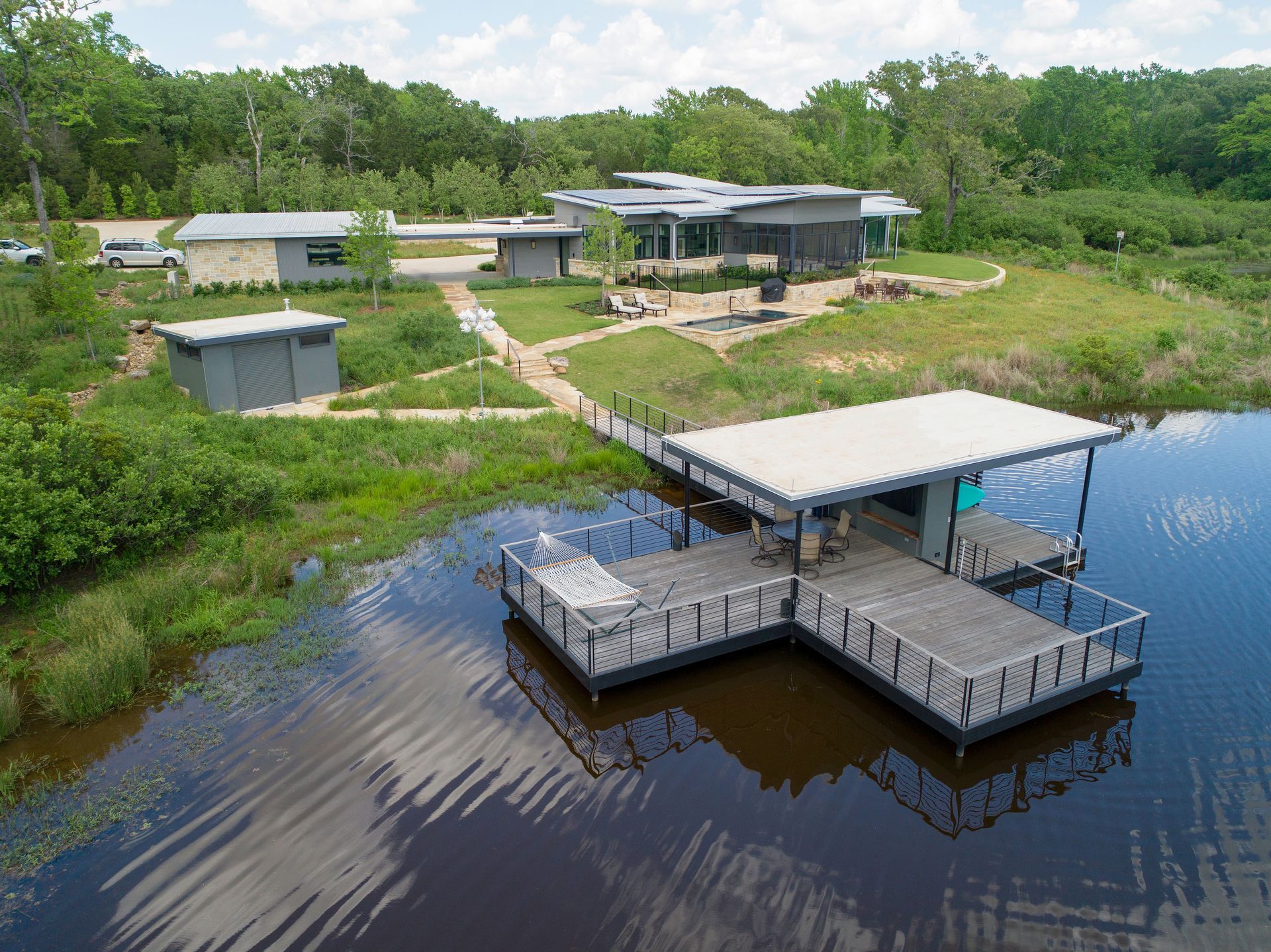 An aerial view shows a modern house and a large, T-shaped floating dock on a lake surrounded by green trees and grass.