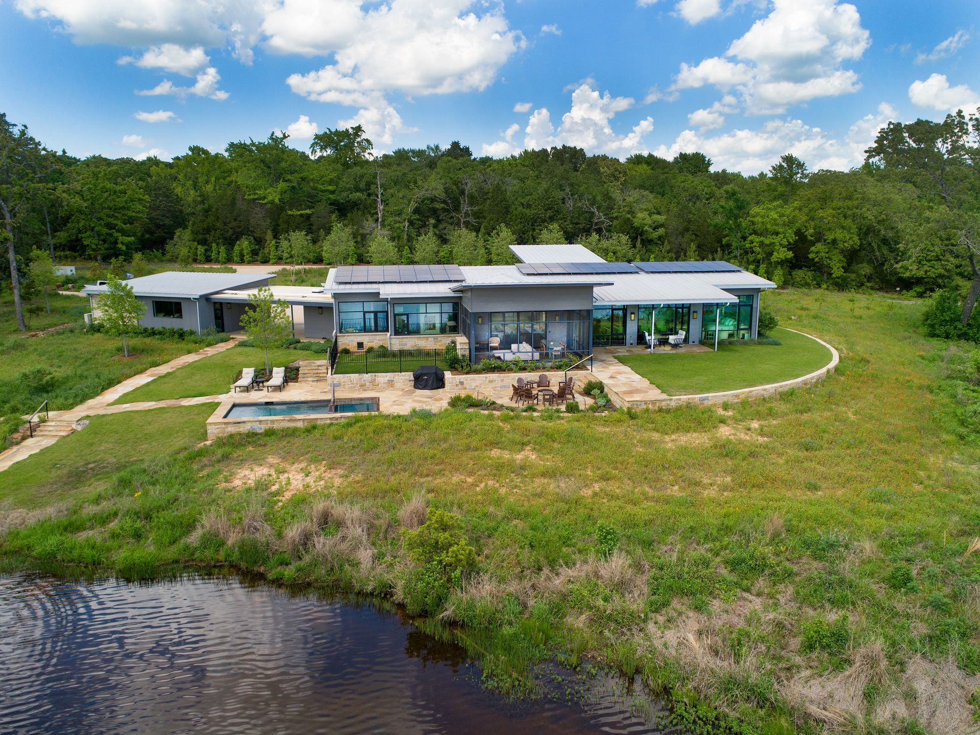 An aerial view of a modern, flat-roofed house set in a grassy field beside a small pond, framed by a forest background.