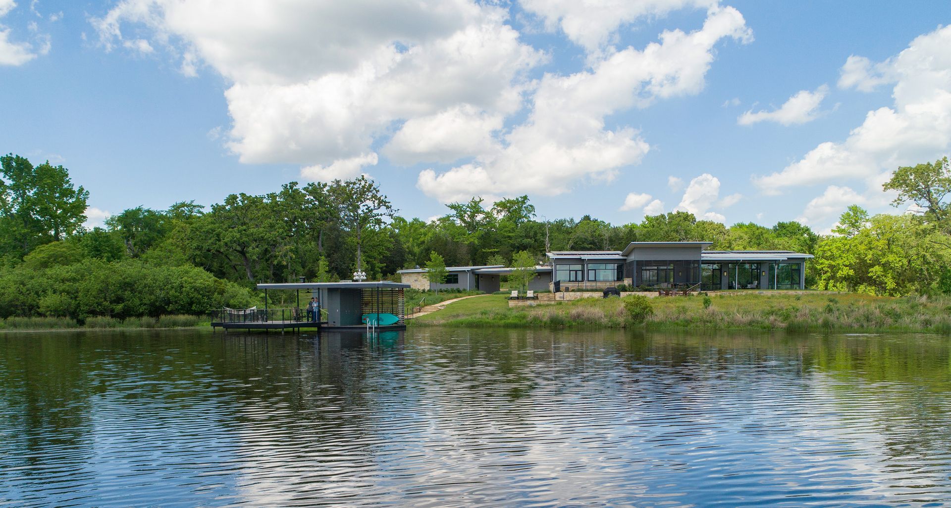 A modern house with floor-to-ceiling windows sits on a grassy lakefront beside a boat dock under a bright, cloudy sky.