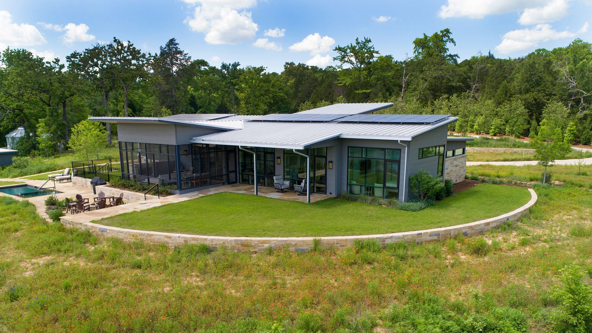 A modern, flat-roofed house with solar panels, surrounded by a stone retaining wall and expansive green lawn and woods.