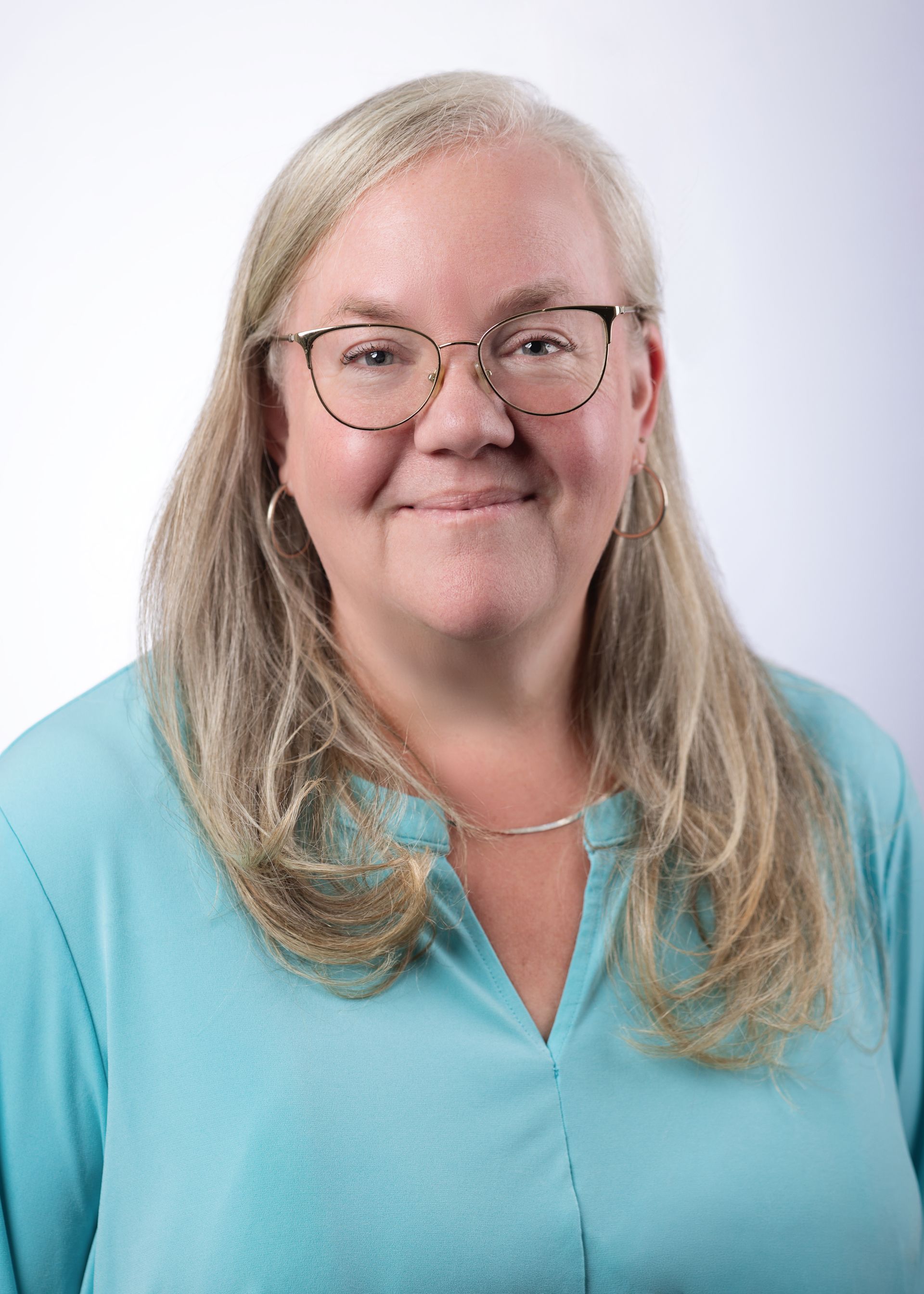 A person with long blonde hair and glasses wearing a light blue top, posed against a plain white background.