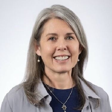 A professional headshot of a smiling person with shoulder-length gray hair, wearing a gray shirt and a necklace.