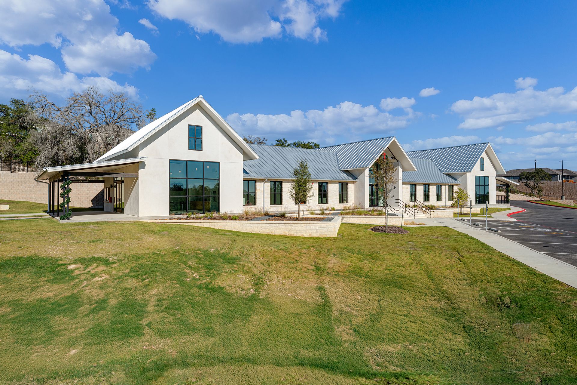 A white, modern, single-story building with a gray metal roof, large windows, and a grassy lawn under a blue sky.