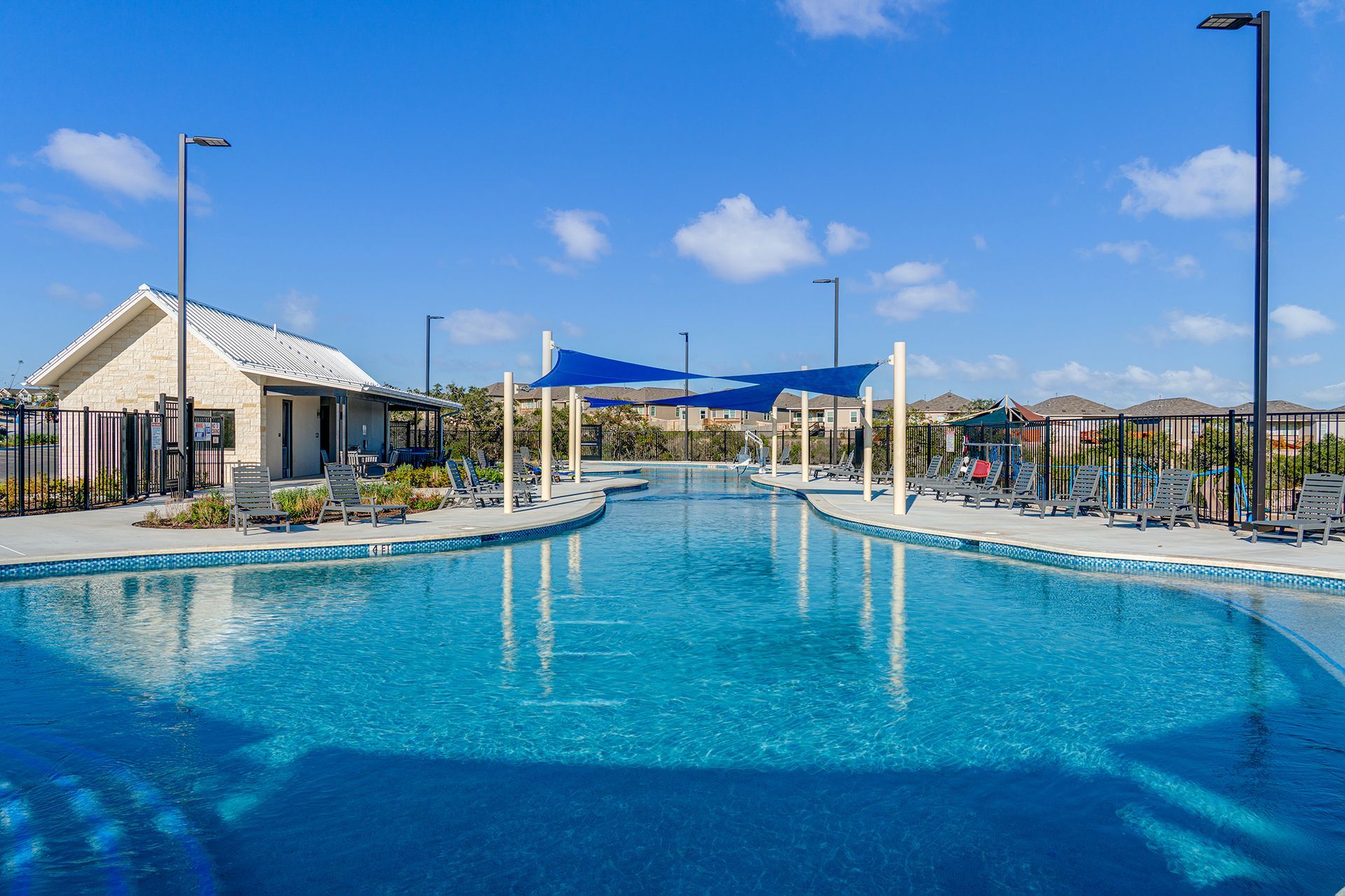 A bright blue swimming pool with a stone building and a shaded lounge area under a clear, sunny sky.