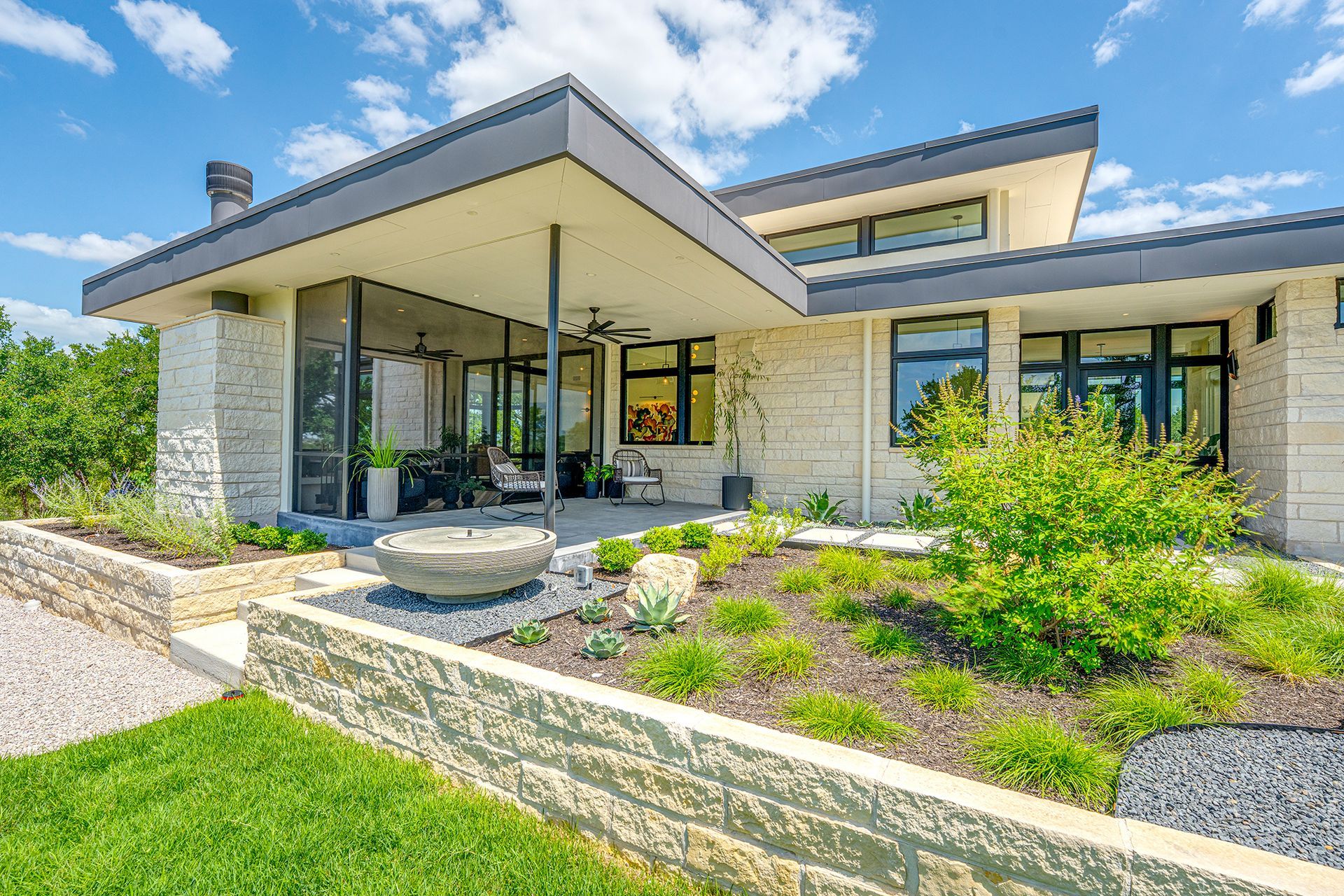 Modern stone house with a covered entry, large windows, and landscaped front yard under a blue sky