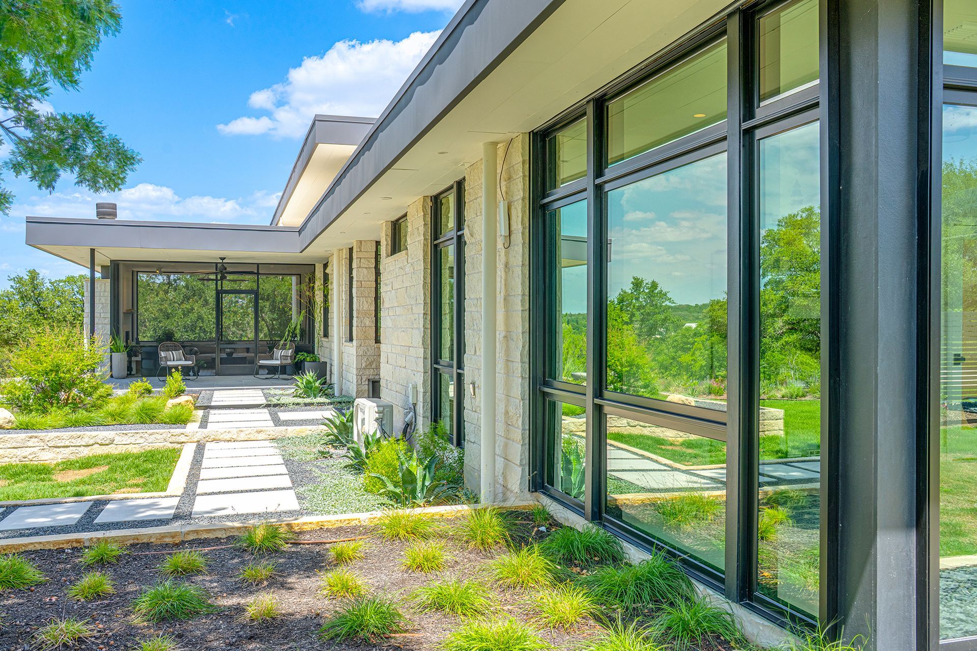 Modern single-story home with a stone exterior, floor-to-ceiling windows, and a stone walkway in a landscaped yard.