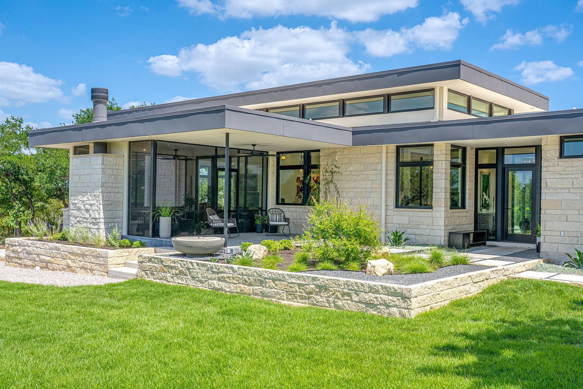 A modern stone home with large windows and a covered patio, surrounded by a lush green lawn and landscaped garden beds.
