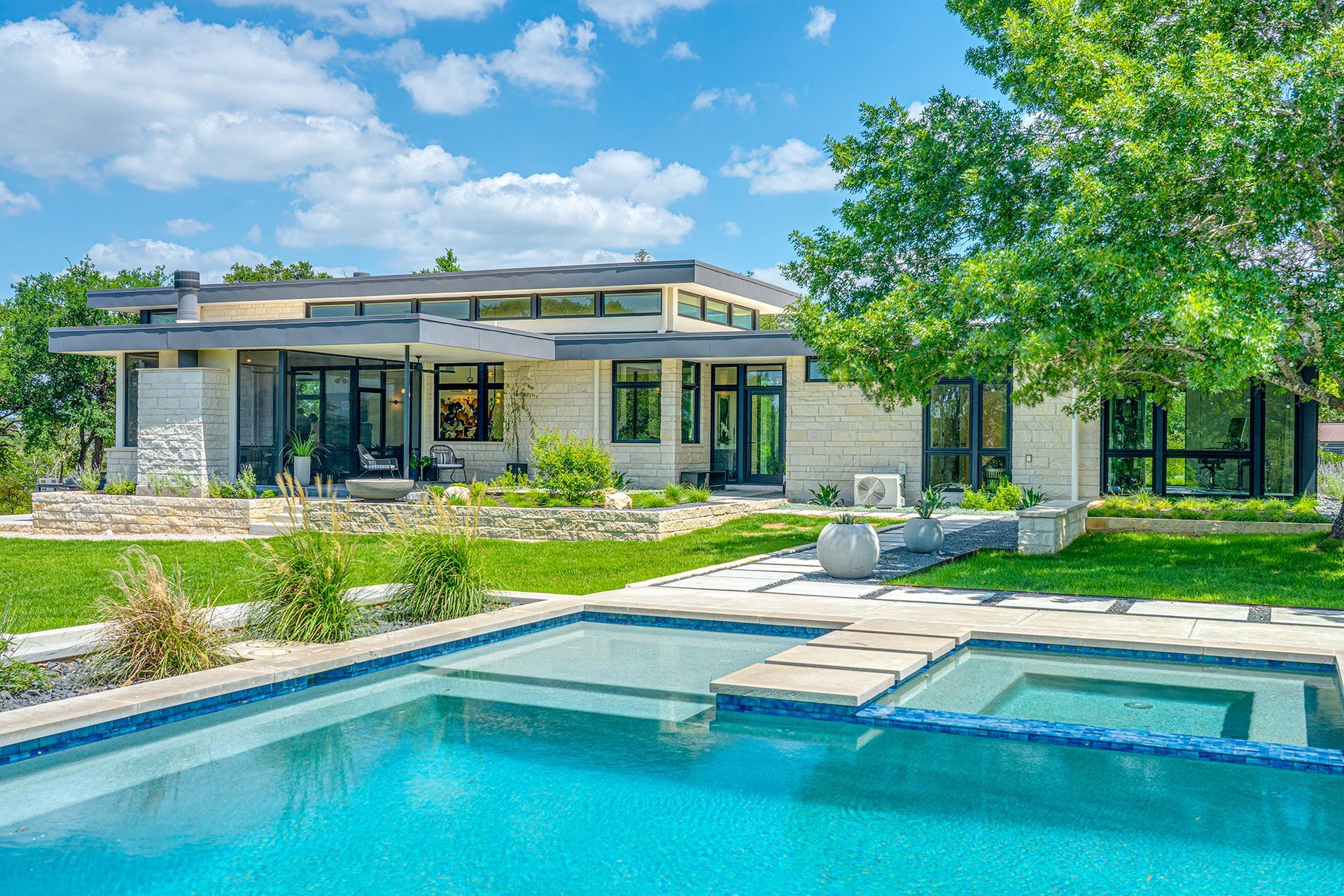 Modern house with a patio and lush landscaping beside a turquoise swimming pool under a blue sky