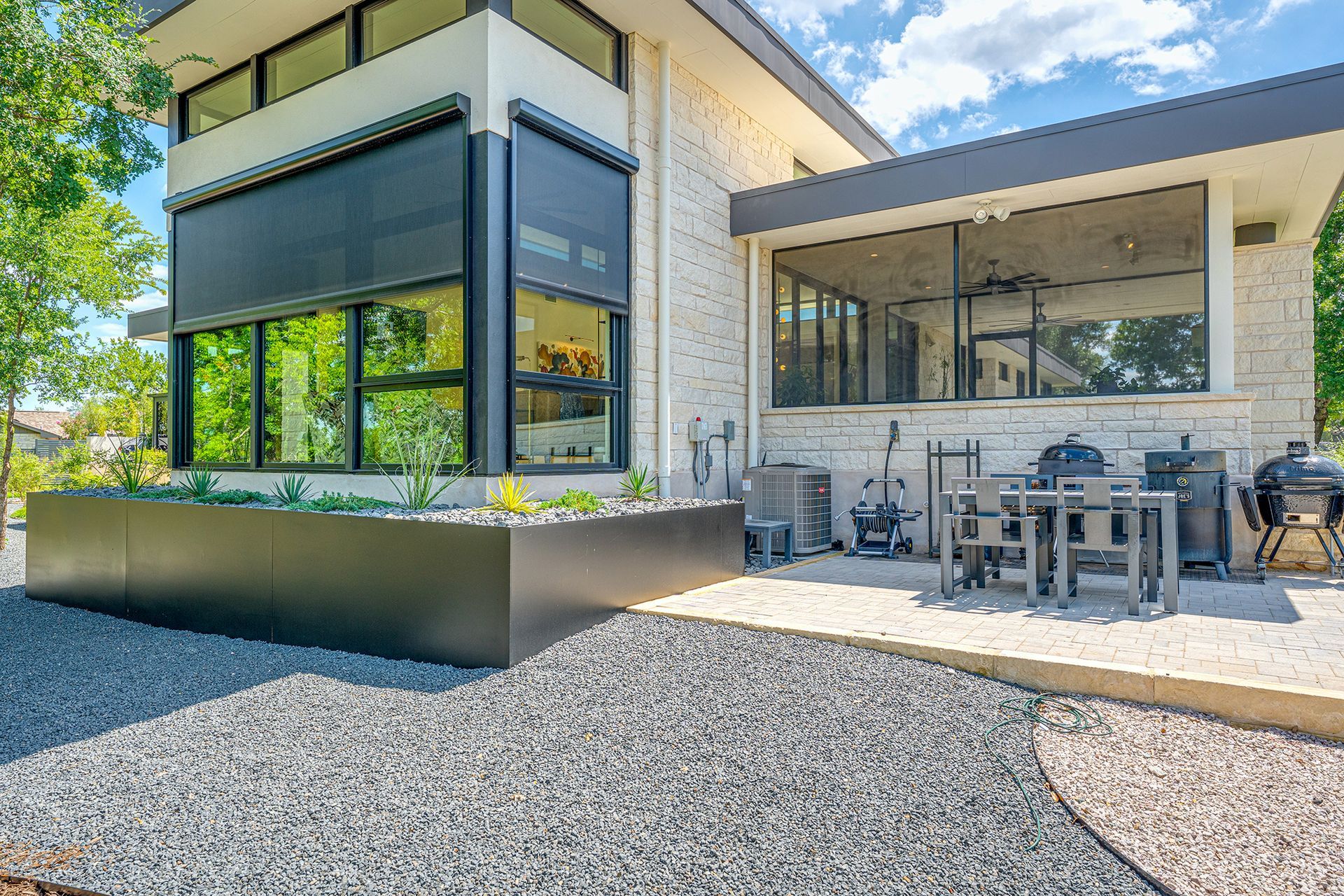 Modern stone house with a black-framed window, gravel yard, raised metal planter, and a patio area with outdoor furniture.