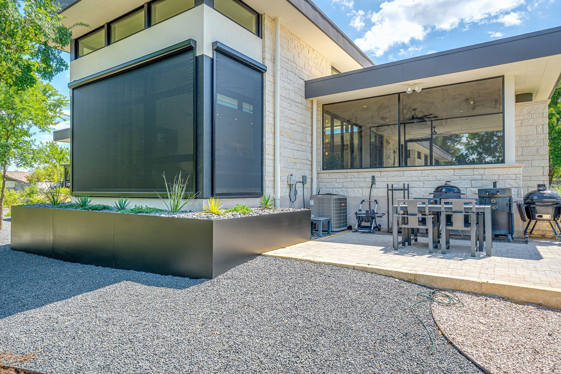 A modern home exterior with a dark-framed patio, metal garden planter, and a stone-covered ground area under a blue sky.