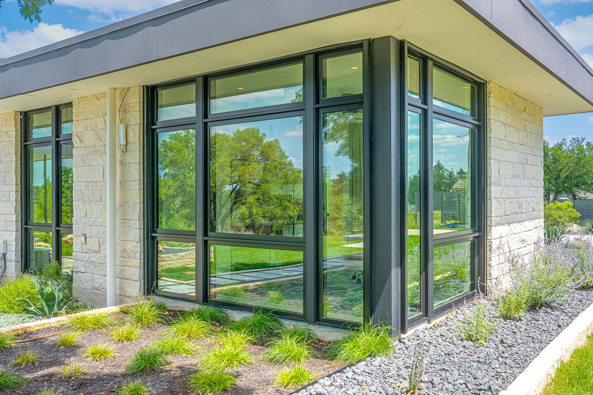 Modern one-story building with cream stone walls, dark metal window frames, and landscaping with gravel and green plants.