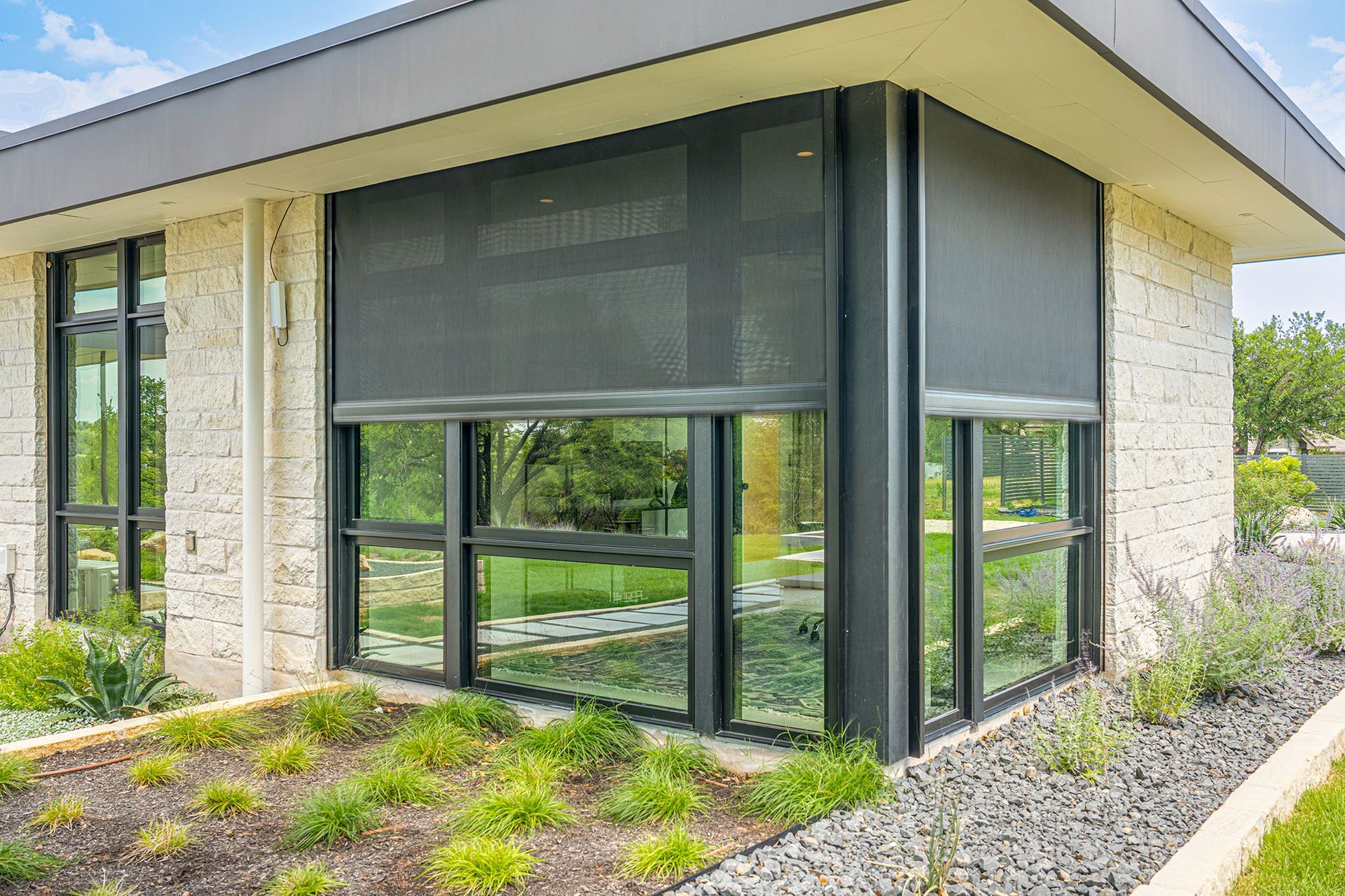 A modern building corner with stone walls and large windows partially covered by dark, retractable sun shades.