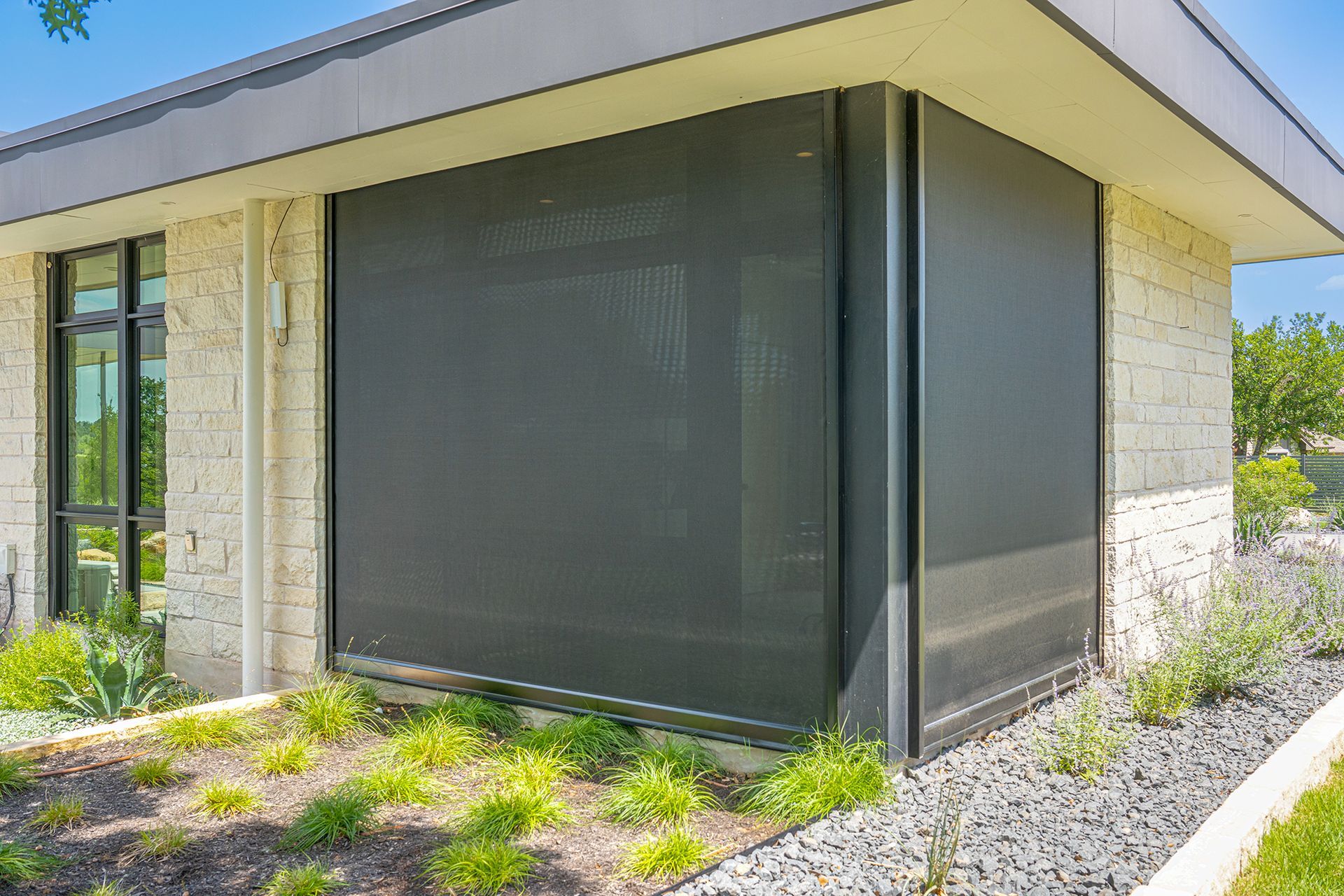 A modern stone building with dark exterior privacy screens installed over large windows, surrounded by landscaping.
