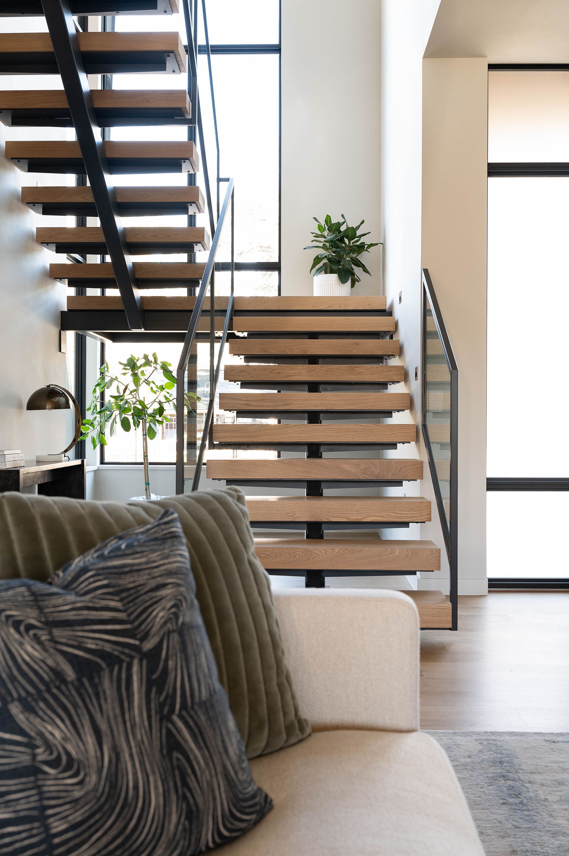 A modern living room features a floating wooden staircase and large windows, viewed from behind a sofa with throw pillows.
