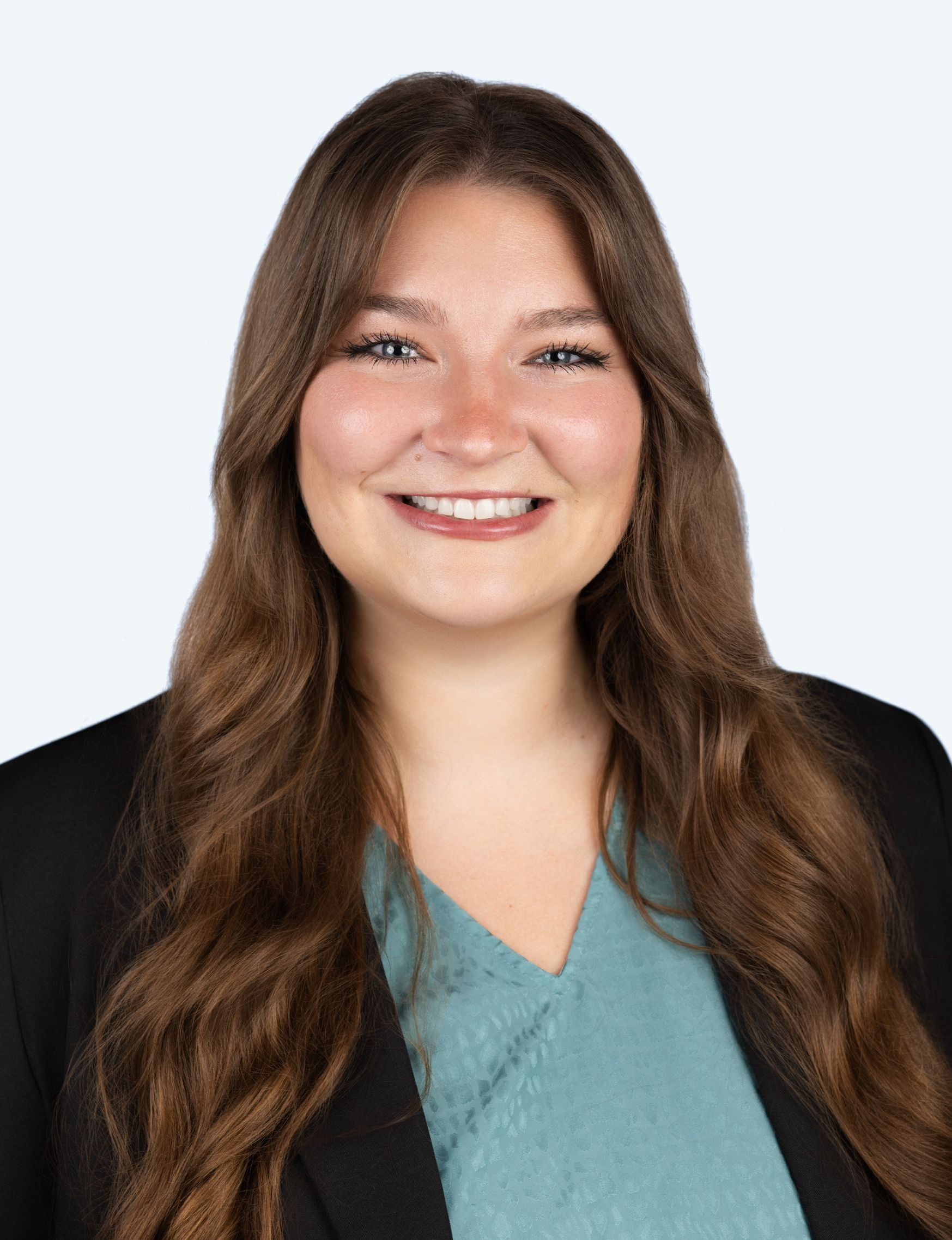 A smiling person with long brown hair wearing a teal top and a black blazer against a light blue background.