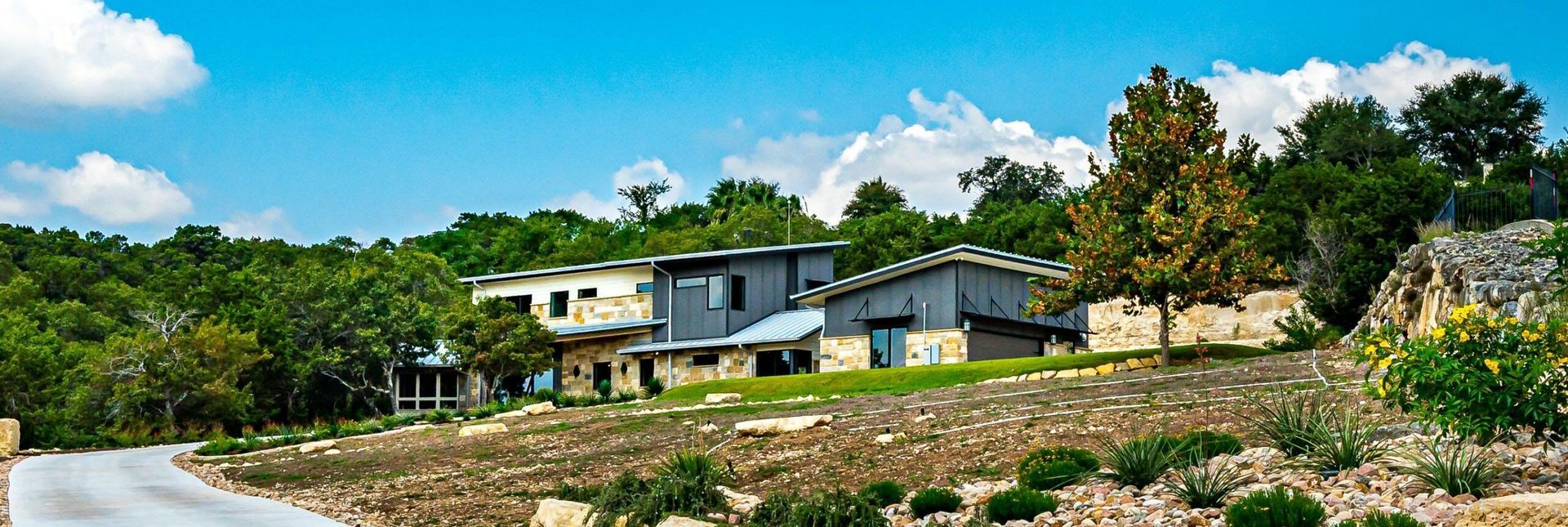 A modern building with a metal roof surrounded by trees and a rocky landscape under a blue sky.