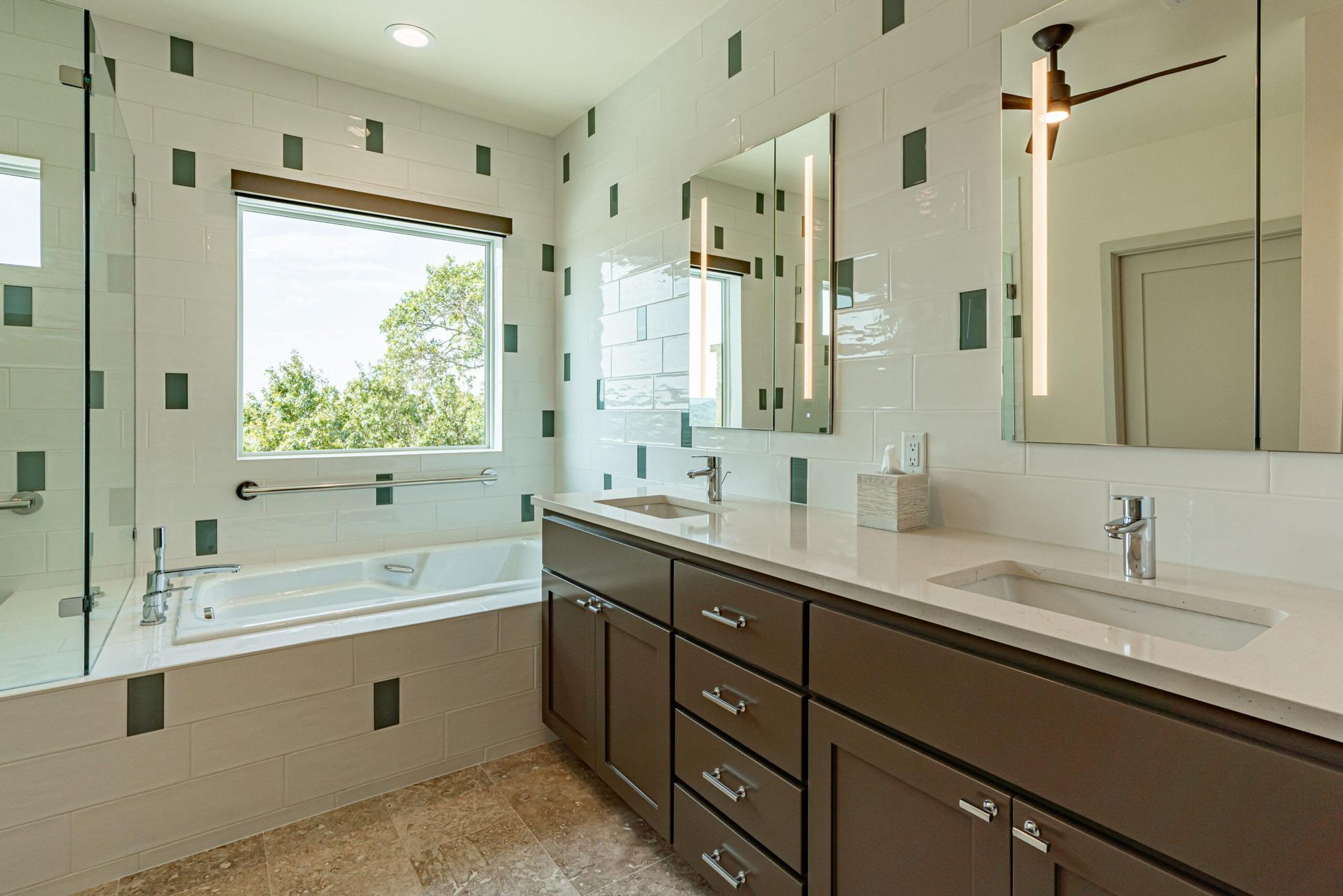 Modern bathroom with a bathtub, dual sinks, white and teal tile, and a large window with a view.