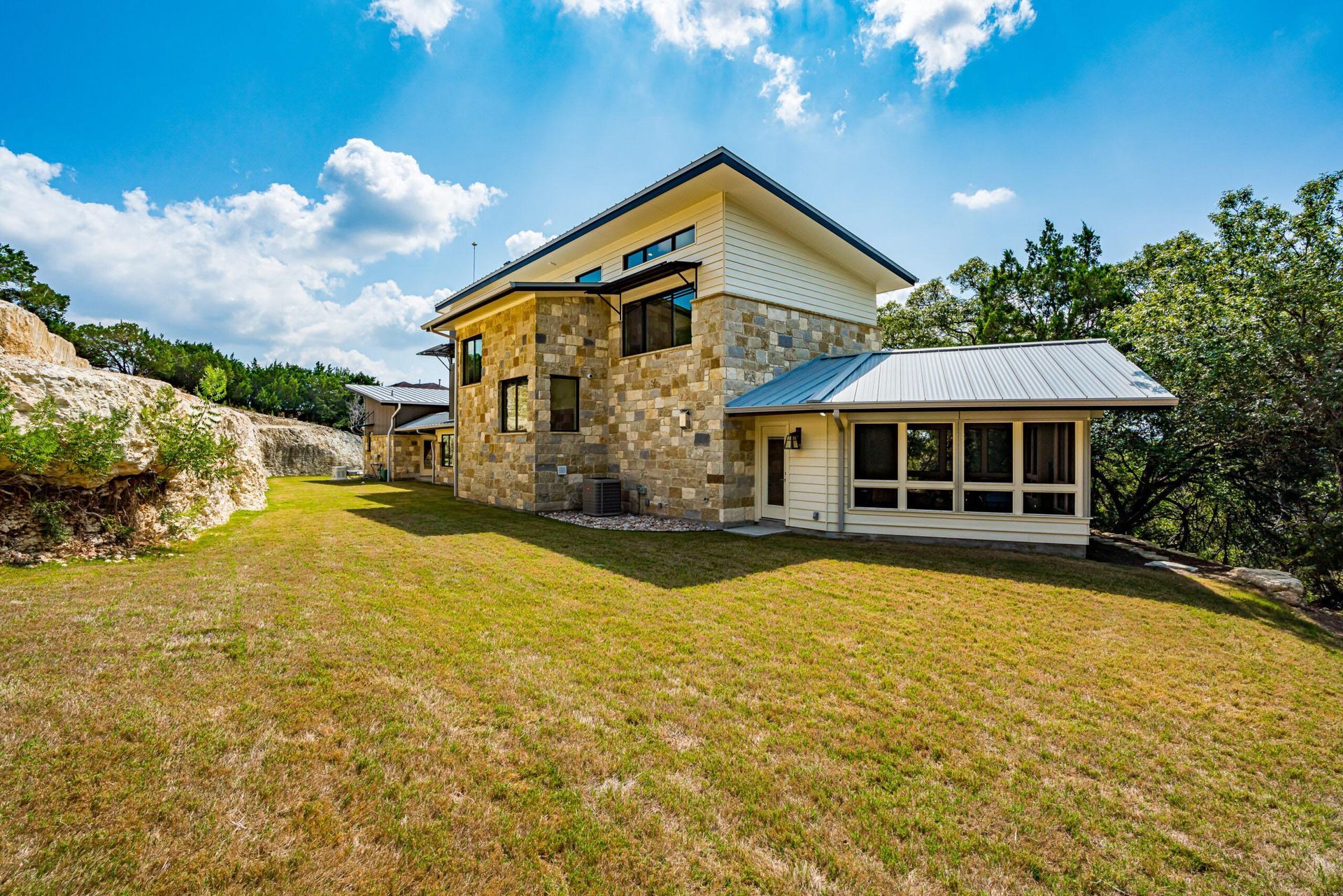 Modern stone house with a screened porch on a grassy hill, bright blue sky.