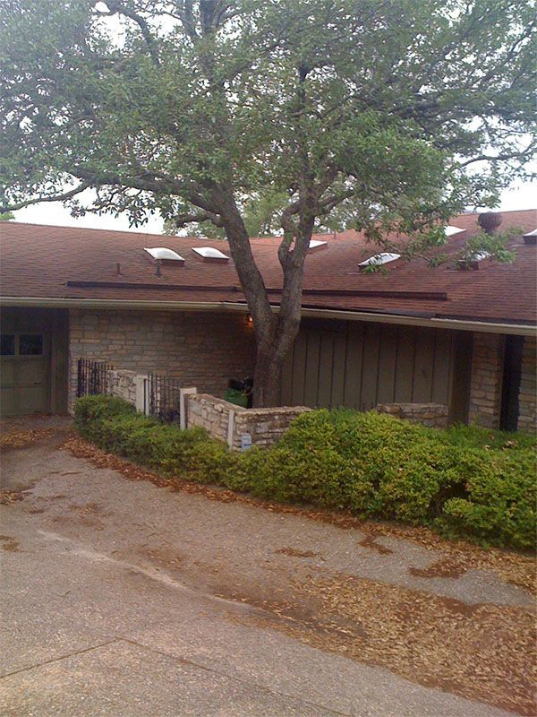 A single-story brick house with a brown roof and a large tree in the front courtyard, seen from a paved driveway.