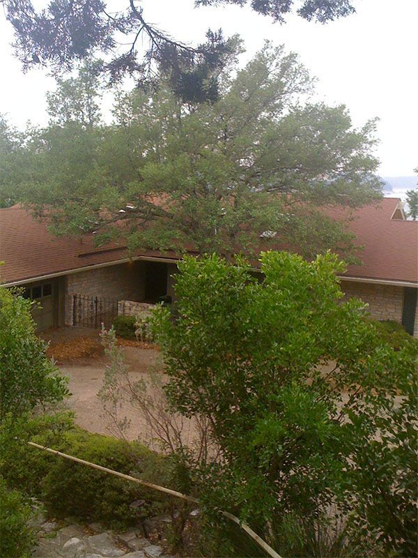 A one-story stone house with a brown shingle roof surrounded by green trees and a driveway.