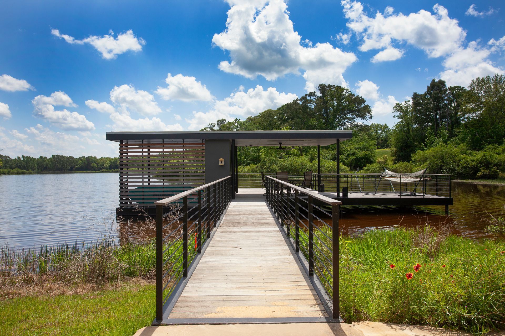 A wooden walkway leads to a modern, covered dock on a calm lake under a bright, cloudy sky.