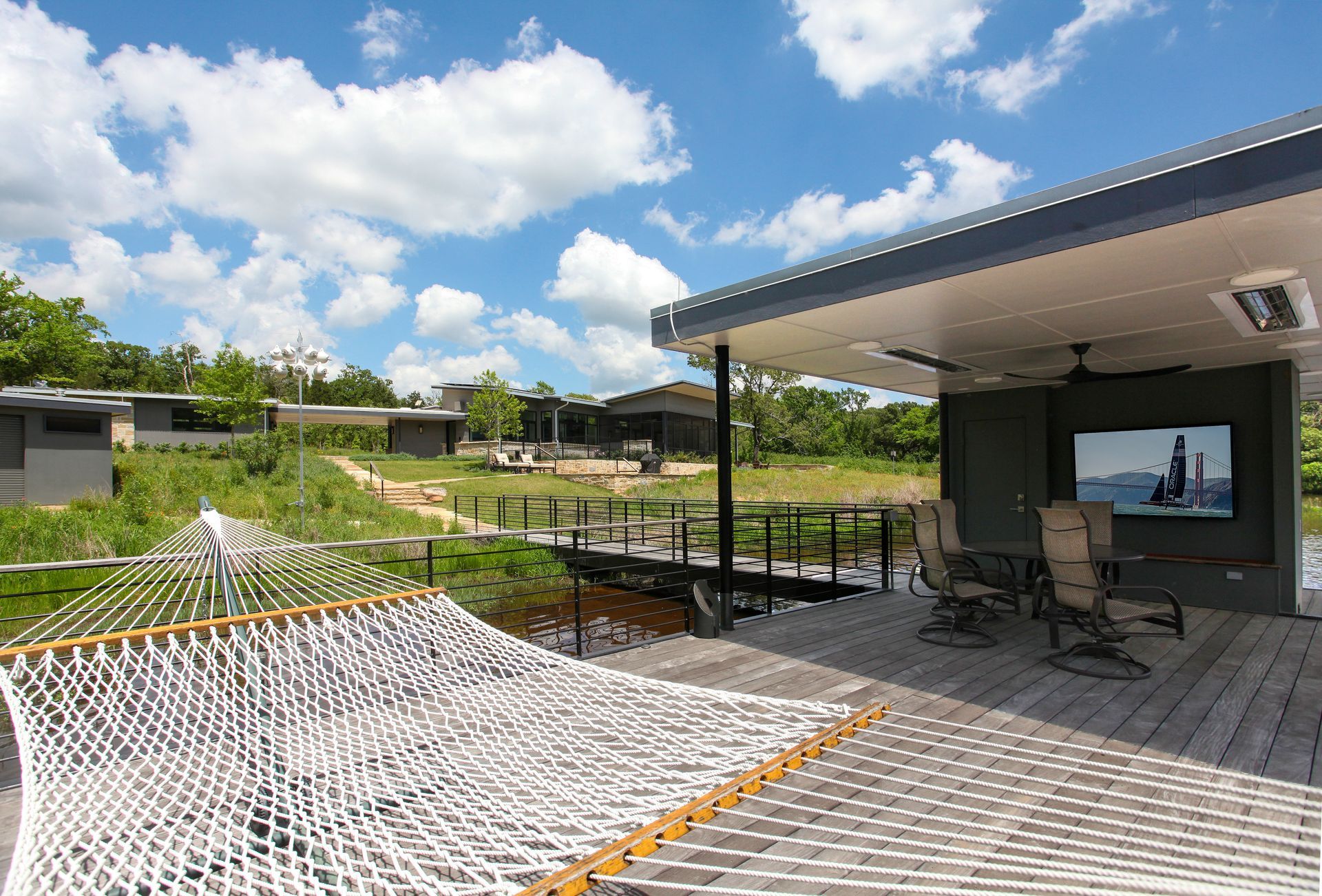 A white rope hammock on a wooden deck overlooks a grassy clearing with modern buildings and a television screen.