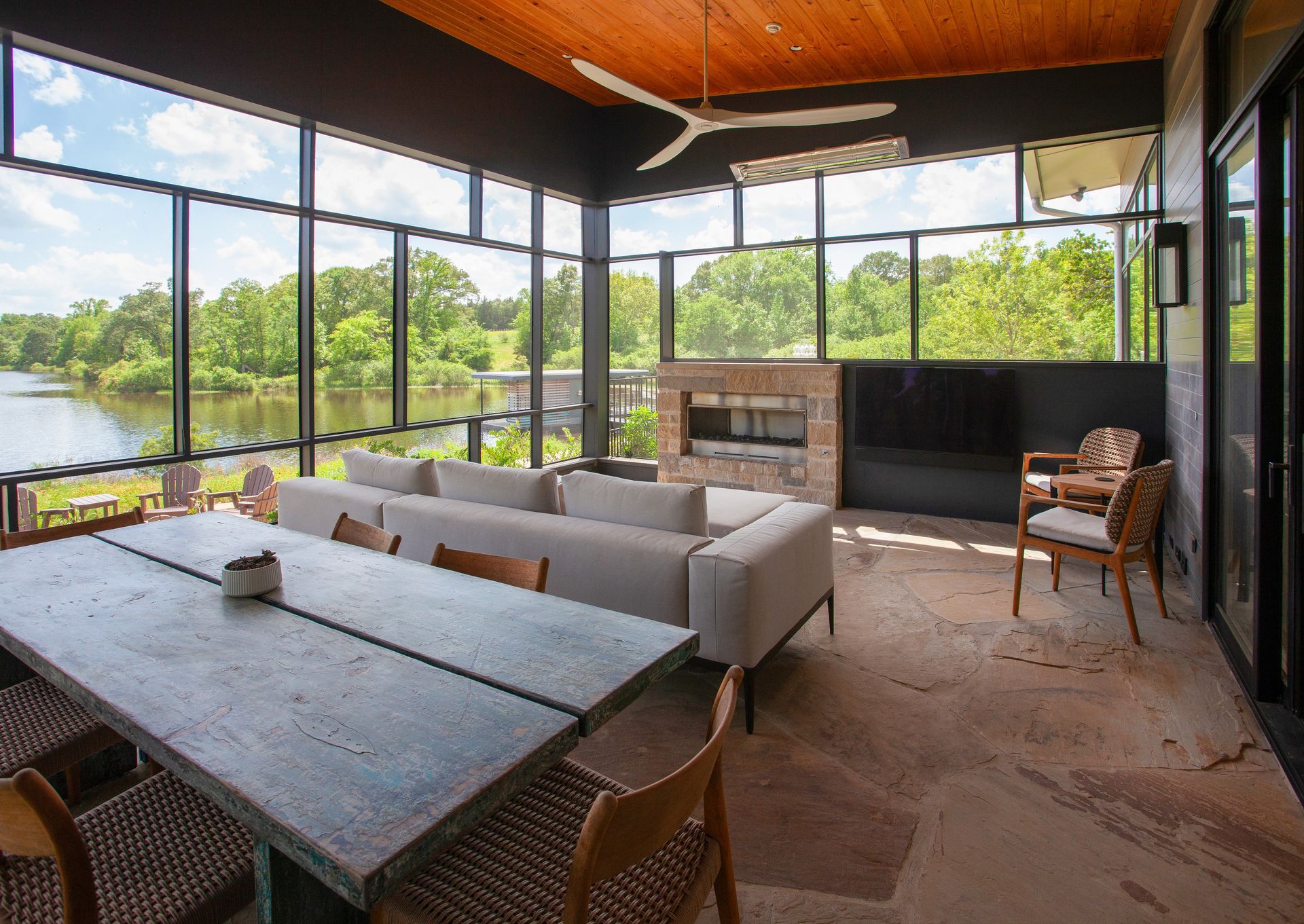 A modern sunroom with a stone floor, grey sofa, concrete dining table, and a fireplace overlooking a river.