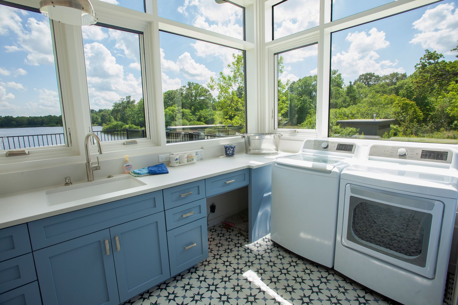 A bright laundry room with blue cabinets, white countertops, a sink, and white appliances, overlooking a lake and trees.
